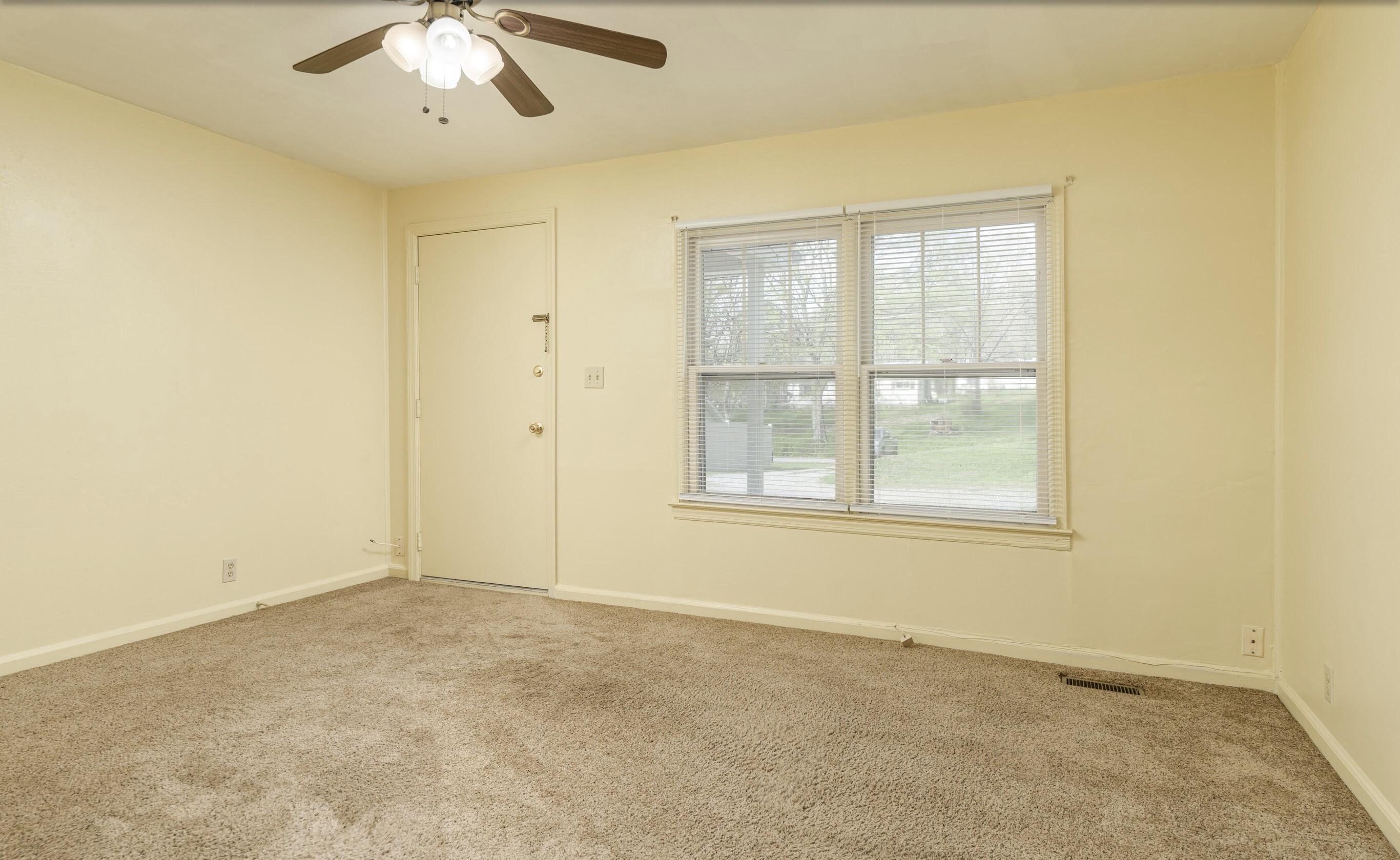Empty carpeted room with a ceiling fan and a large window with blinds.