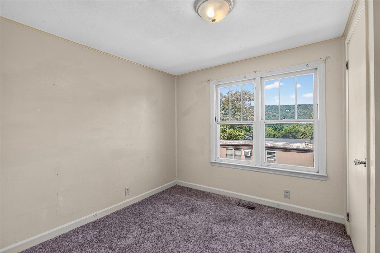 Empty bedroom with gray-purple carpet and twin windows overlooking a wooded hillside.