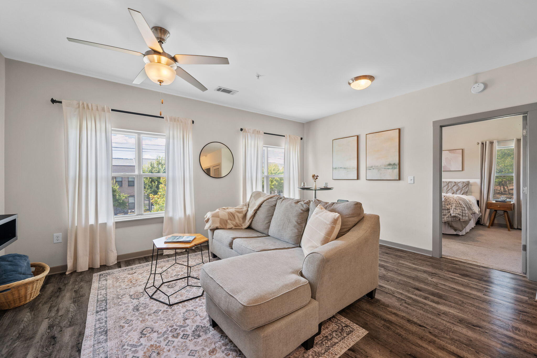 Furnished living room with a beige sectional, patterned rug, and curtained windows; bedroom visible through doorway.