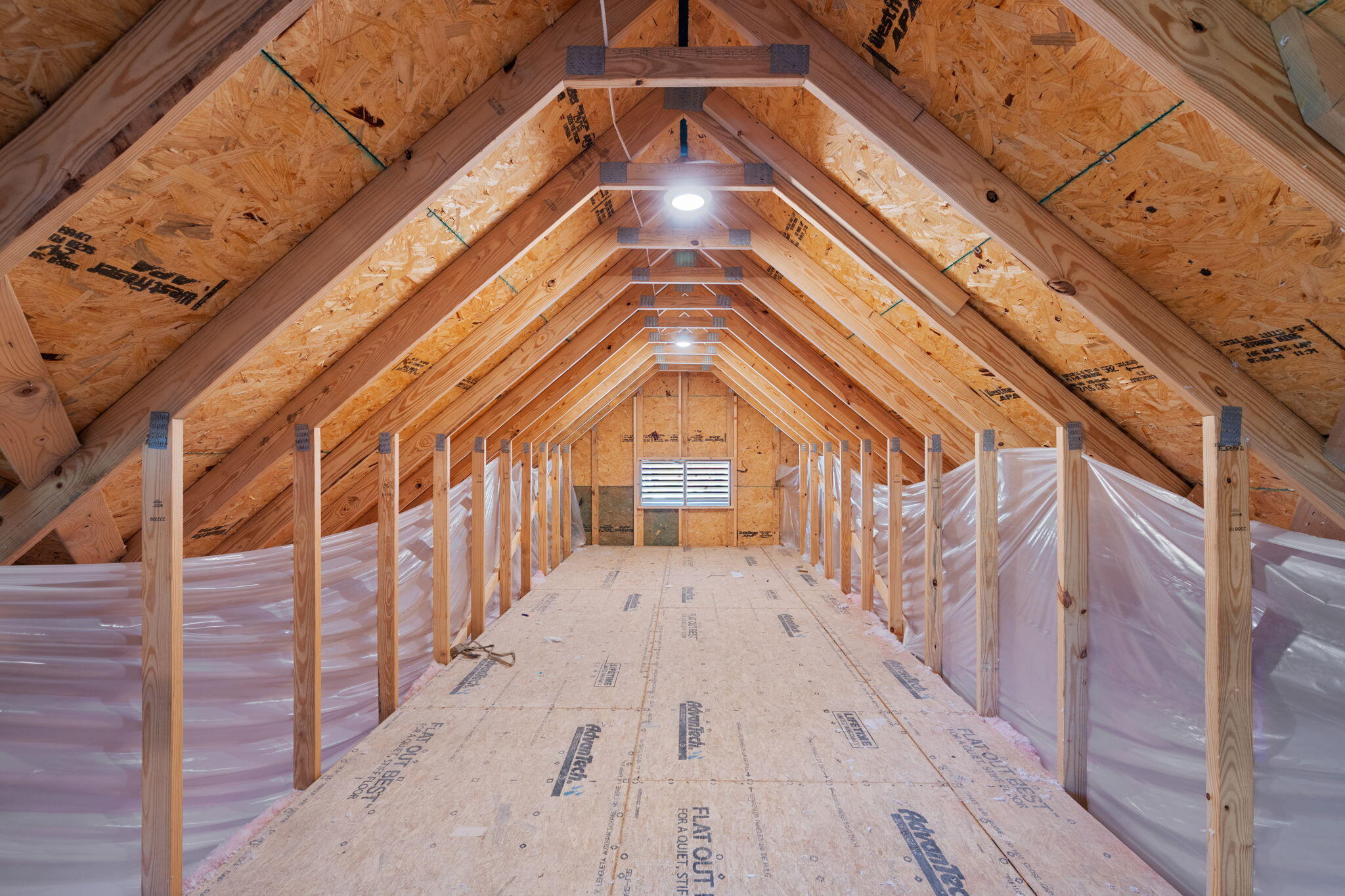 Unfinished attic with exposed roof trusses, plastic-covered sides, and a vent at the far end.