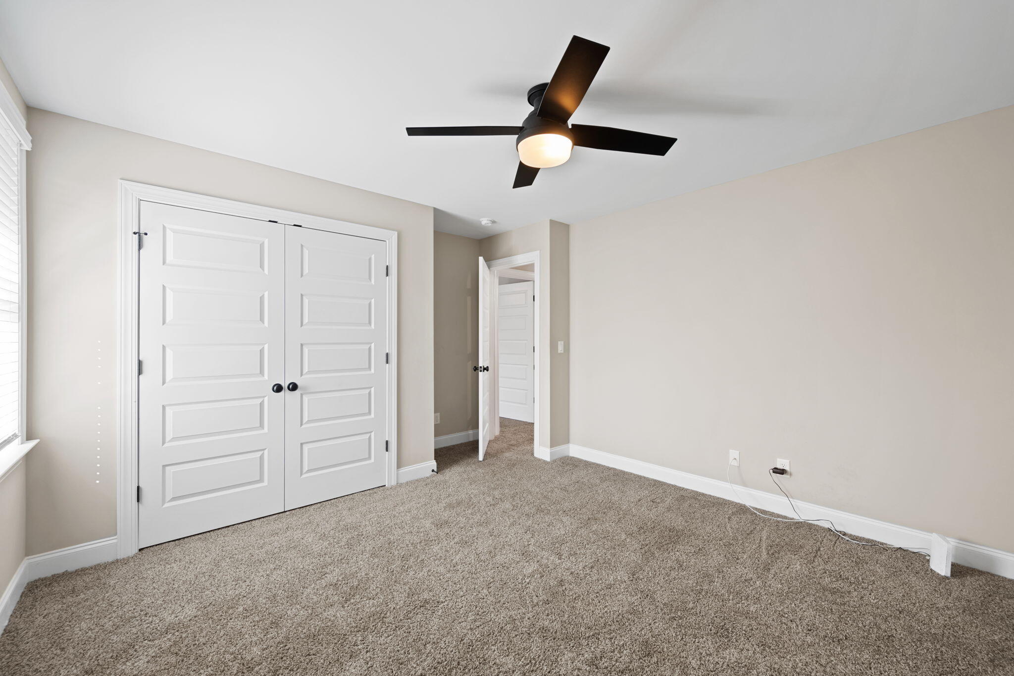 Empty bedroom with white double closet doors, a black ceiling fan, and taupe carpet.