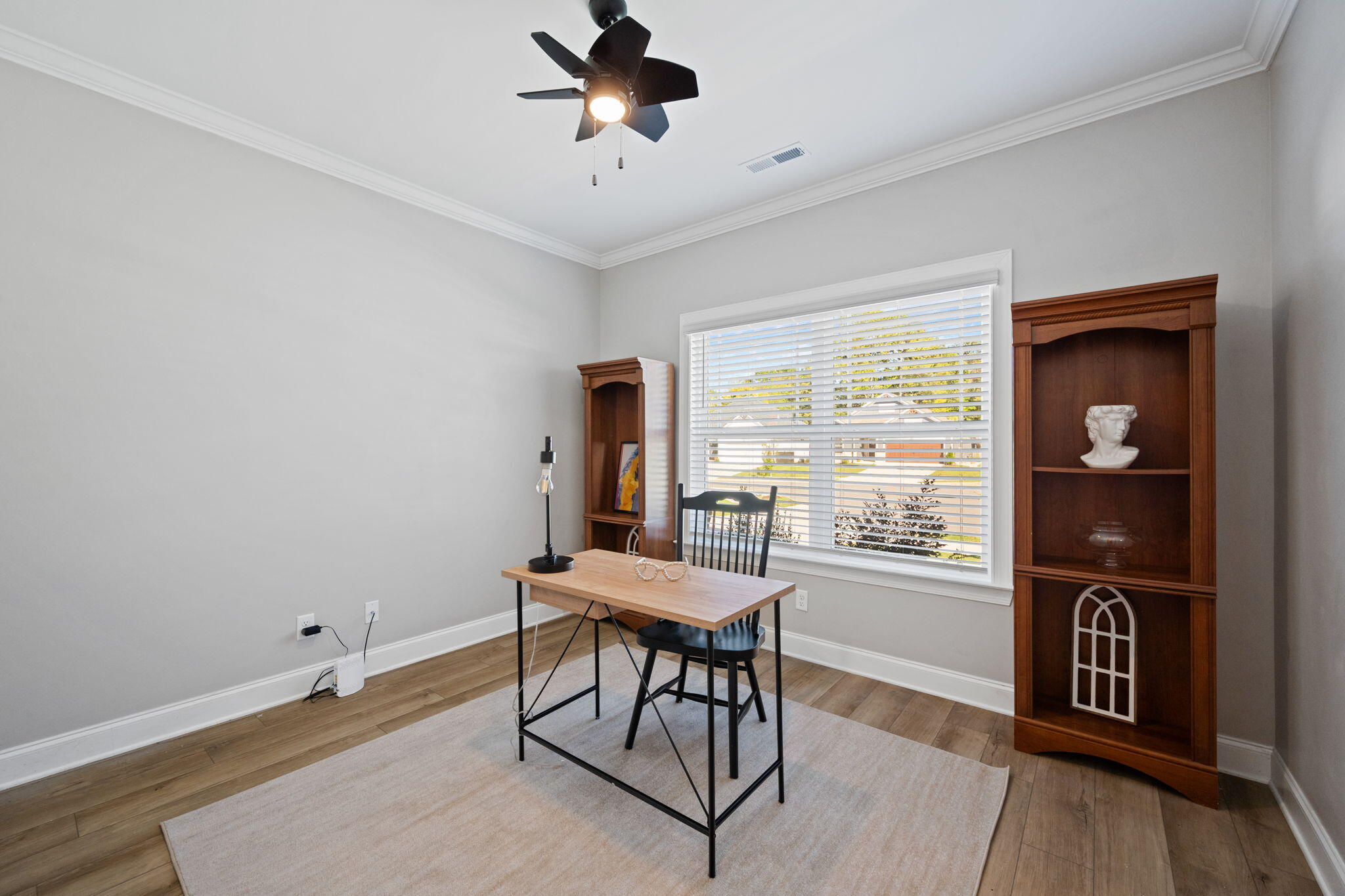 Home office with a light wood desk, wooden bookcases, and a large window with white blinds.
