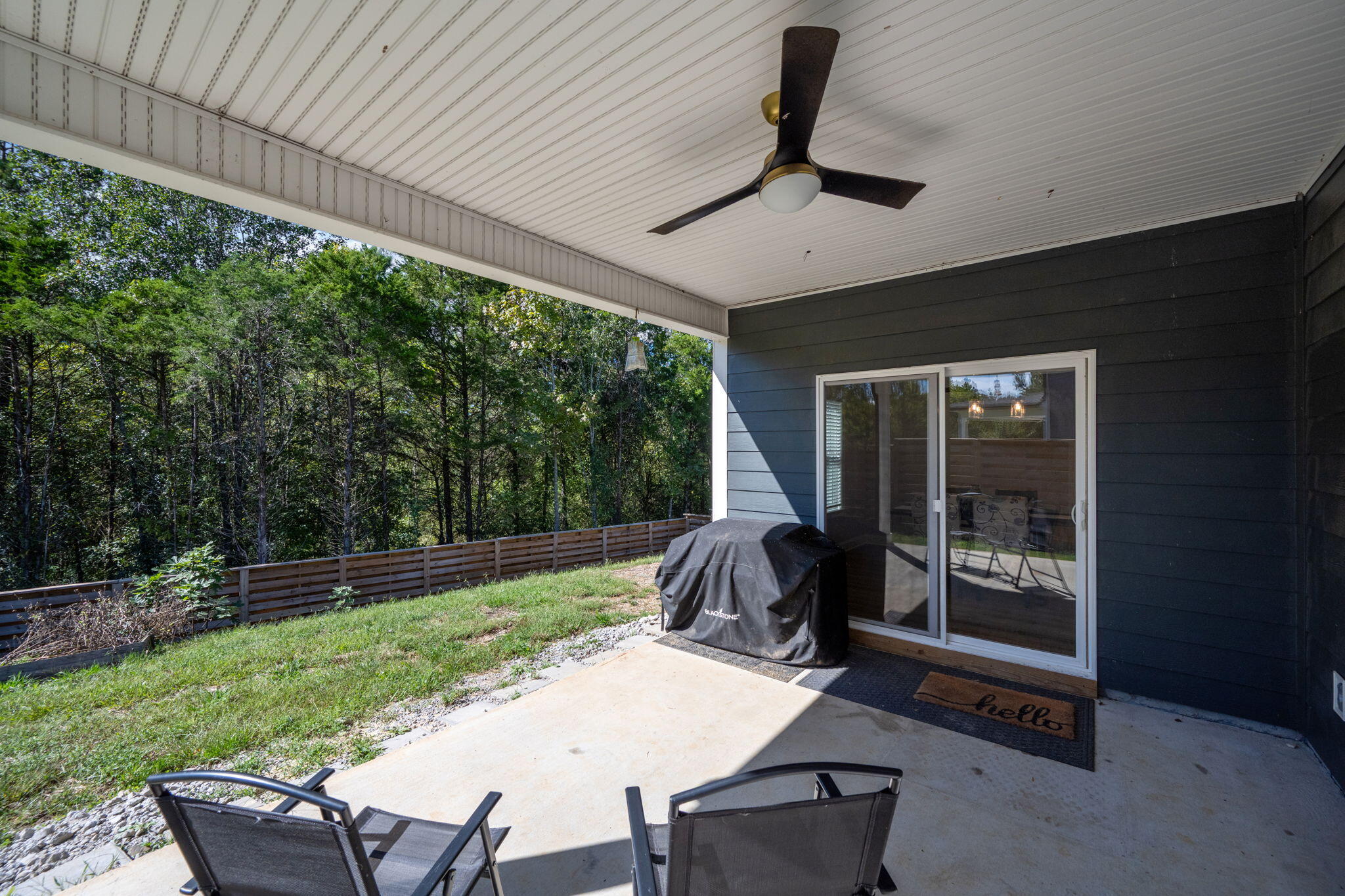 Covered patio with a ceiling fan, grill, and chairs overlooking a fenced, wooded yard.