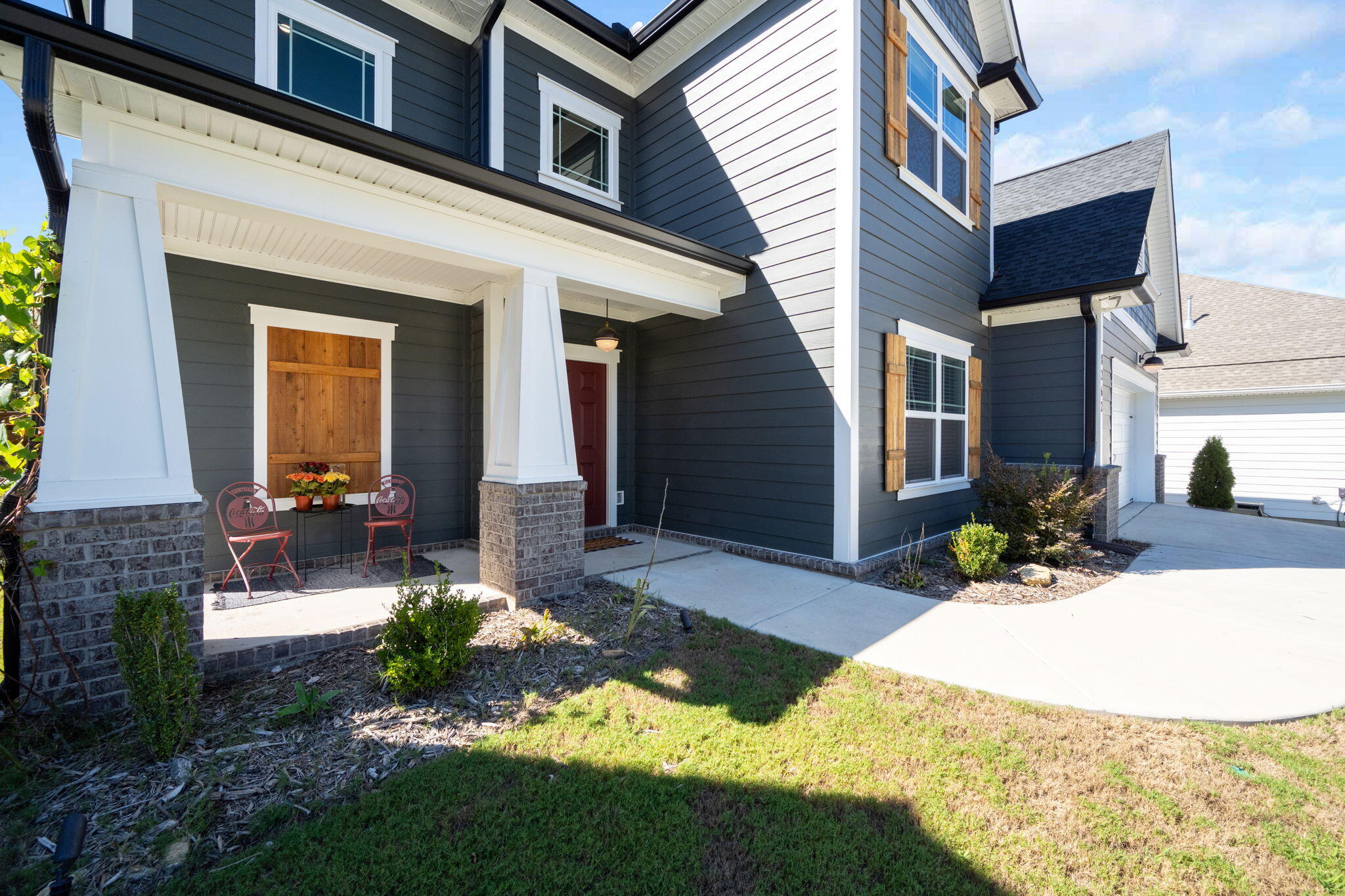 Two-story blue-gray home with a covered porch, red door, wooden shutters, and red metal chairs.