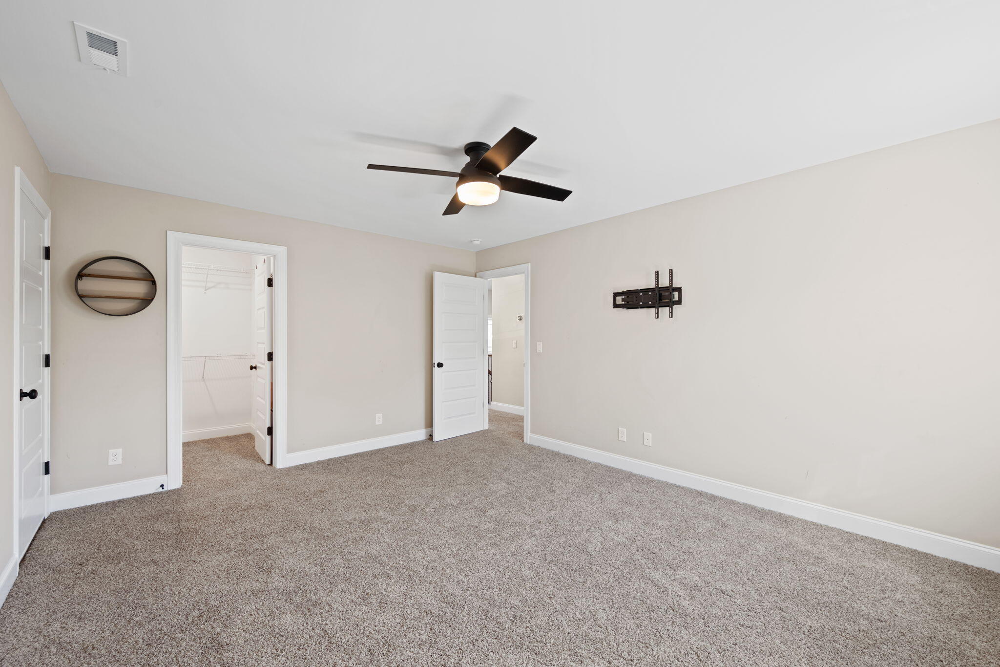 Empty carpeted bedroom with a ceiling fan, TV mount, and open wire-shelf closet.