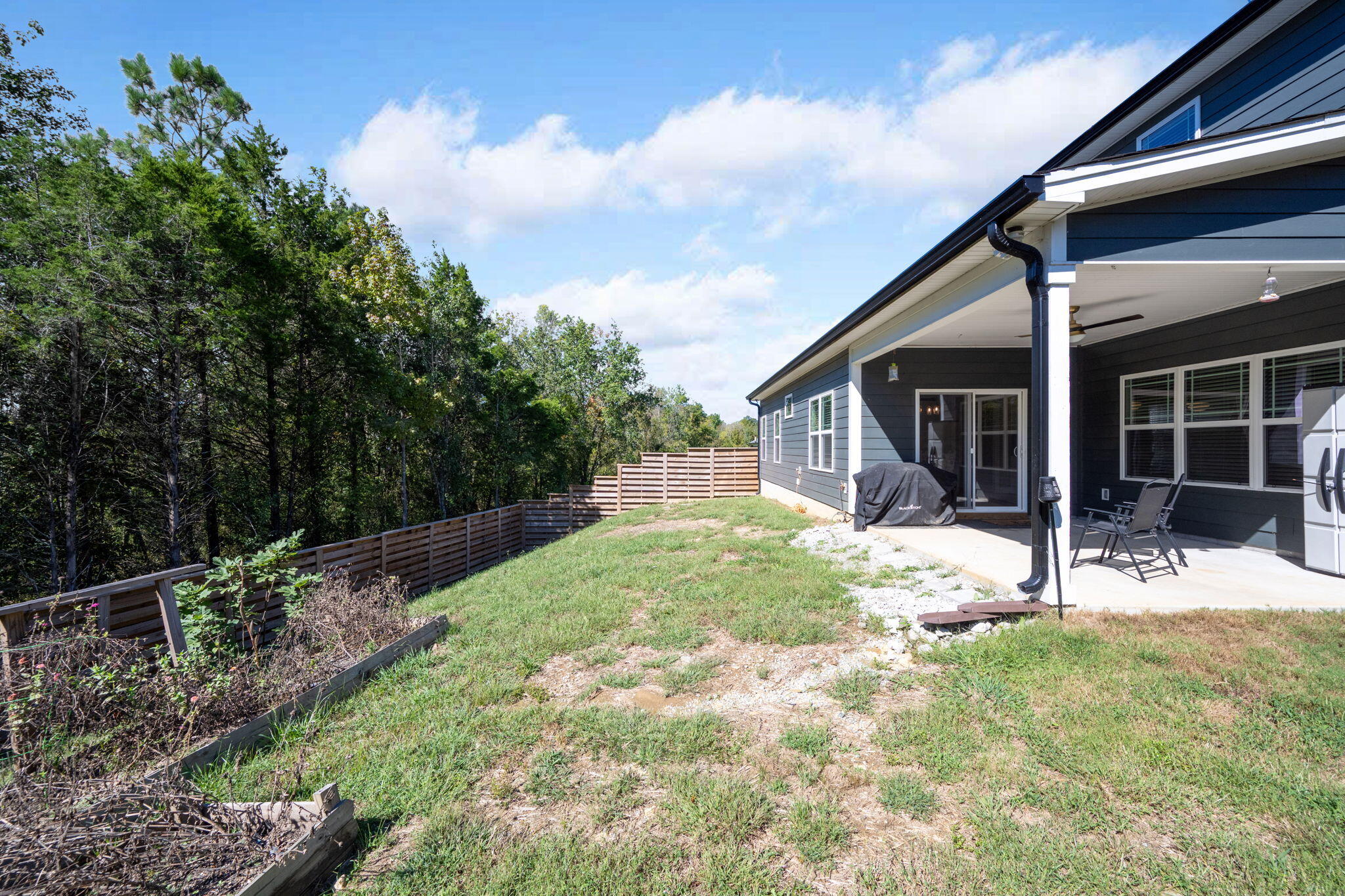 Backyard with covered patio, outdoor seating, grassy yard, and wooden fence beside dense trees.