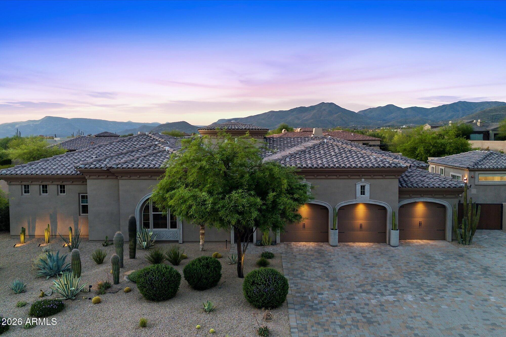 Desert-style home with three lit garage doors, drought-tolerant landscaping, and mountains behind it at dusk.