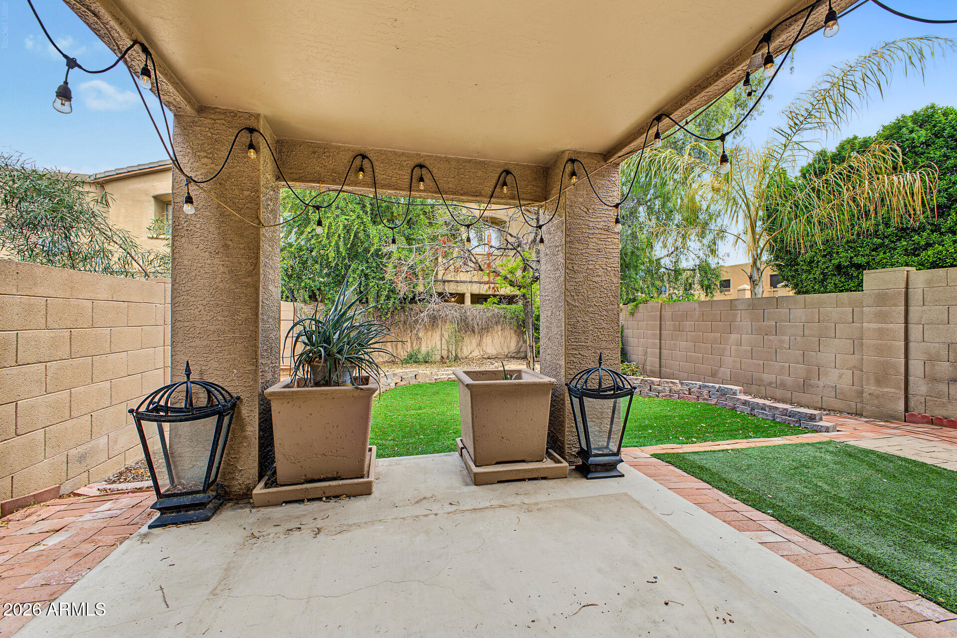 Covered patio with string lights, planters, and lanterns beside a walled backyard lawn.