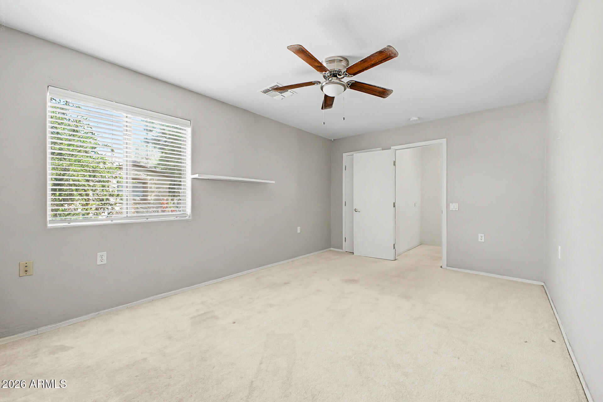 Empty bedroom with beige carpet, a large window with blinds, and a wood-blade ceiling fan.