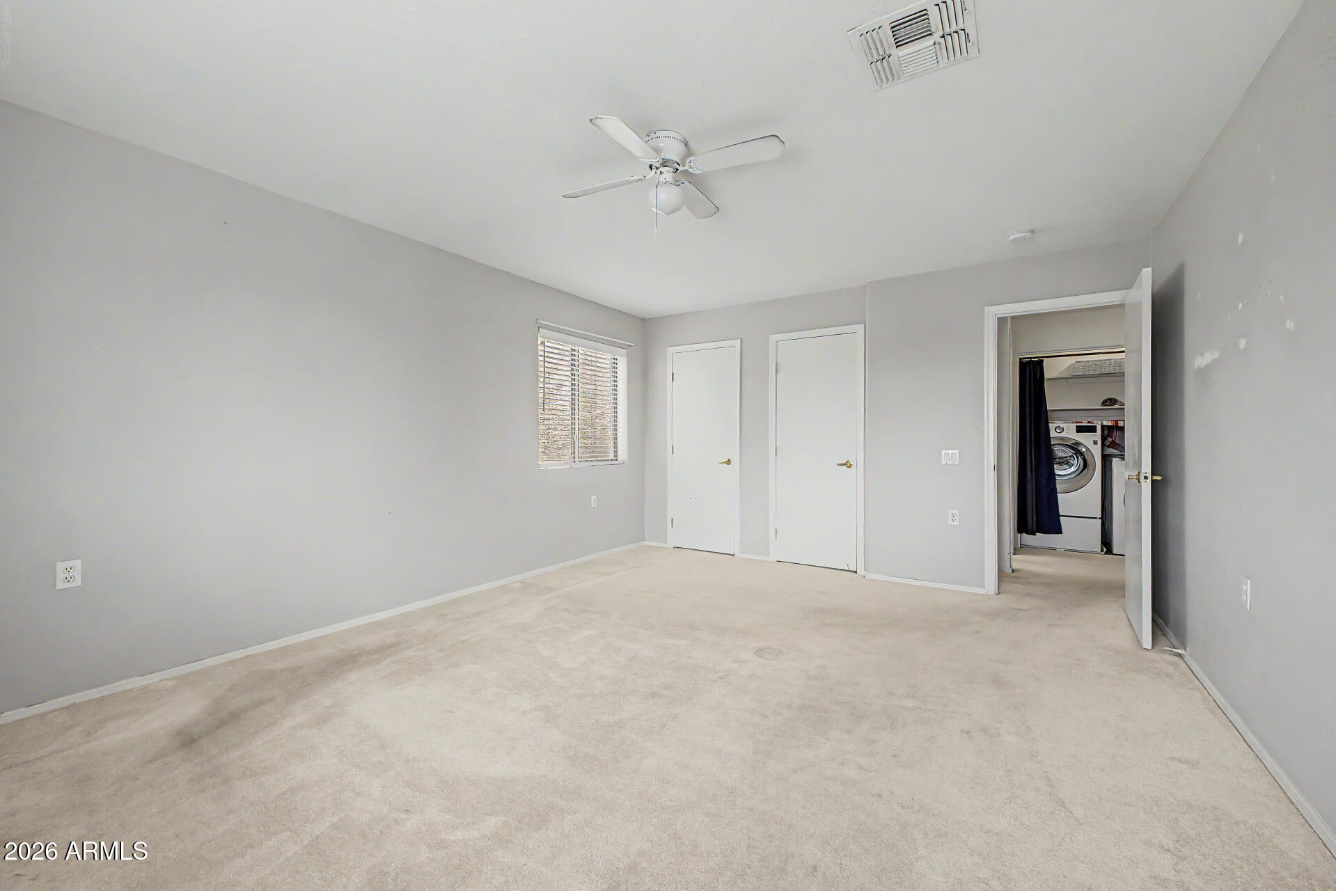 Empty carpeted bedroom with a ceiling fan, window blinds, and laundry area visible through an open doorway.