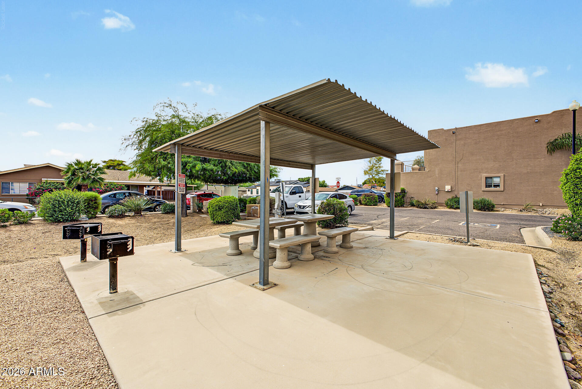 Shared picnic area with concrete tables under a metal canopy and grills nearby.