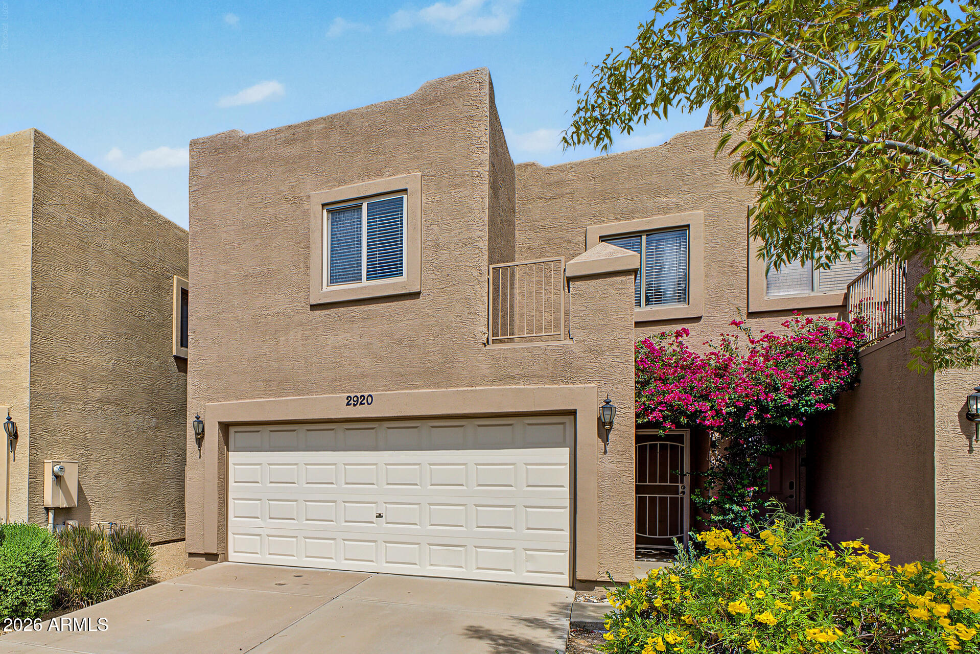 Two-story stucco home with a white double garage and flowering entry garden.