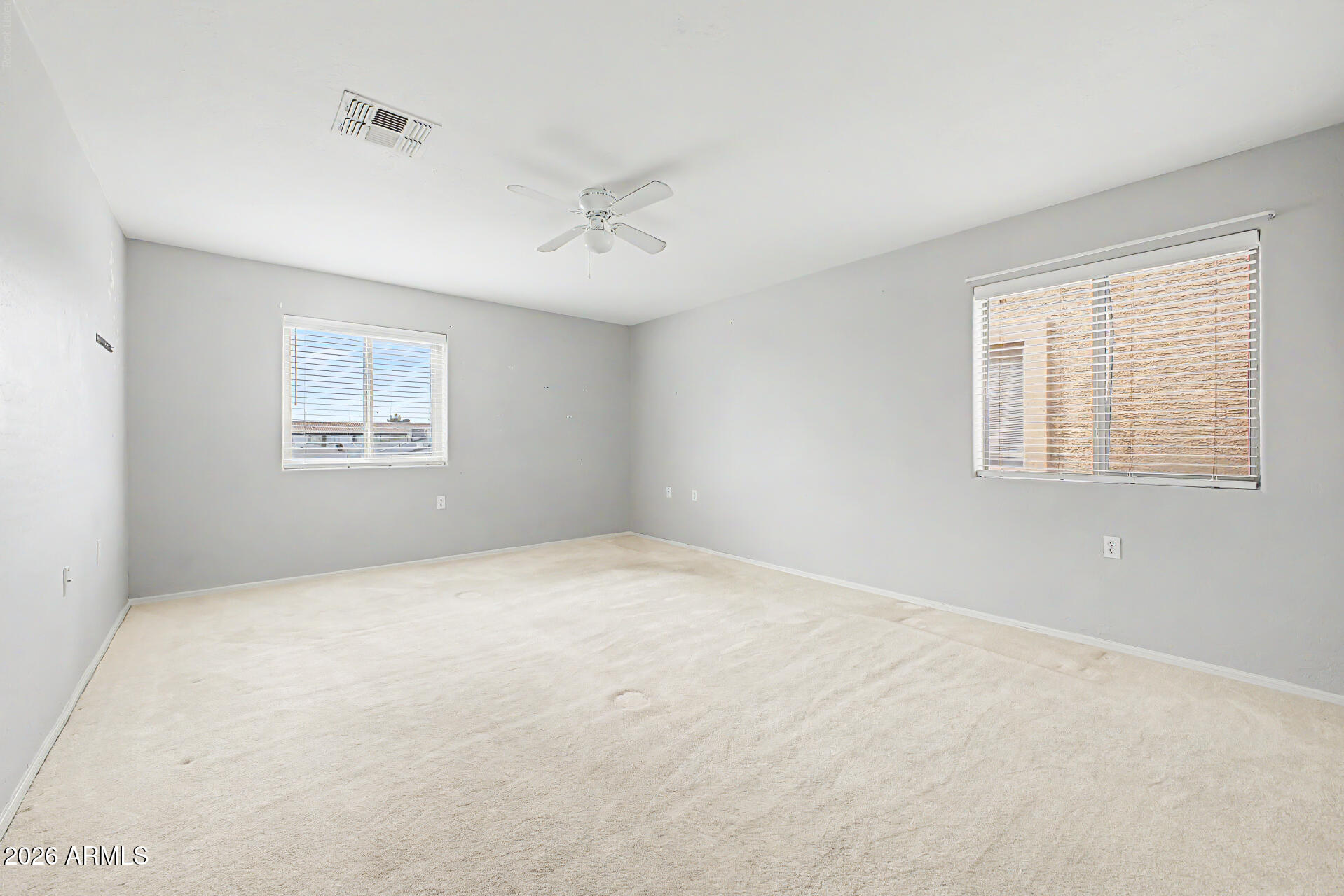 Empty carpeted room with gray walls, two windows with blinds, and a ceiling fan.