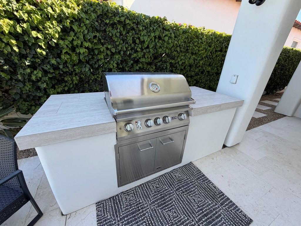 Outdoor kitchen with stainless steel built-in grill, pale stone counter, and tall hedge.