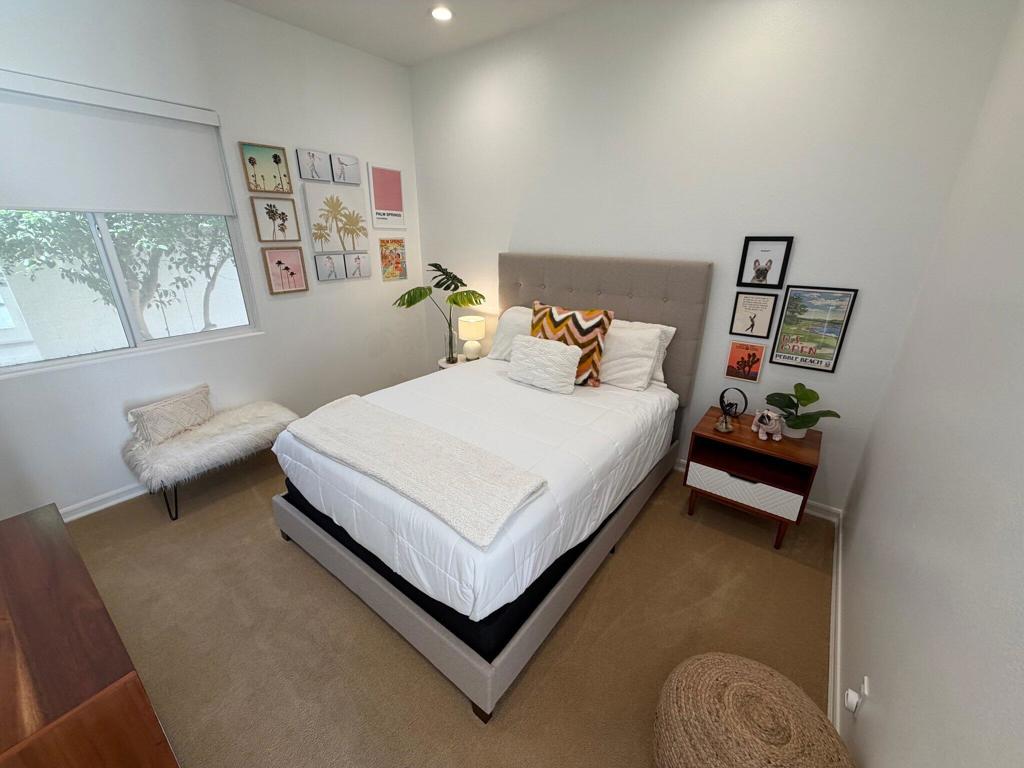 Staged bedroom with a white bed, colorful accent pillows, and framed prints beside a window.