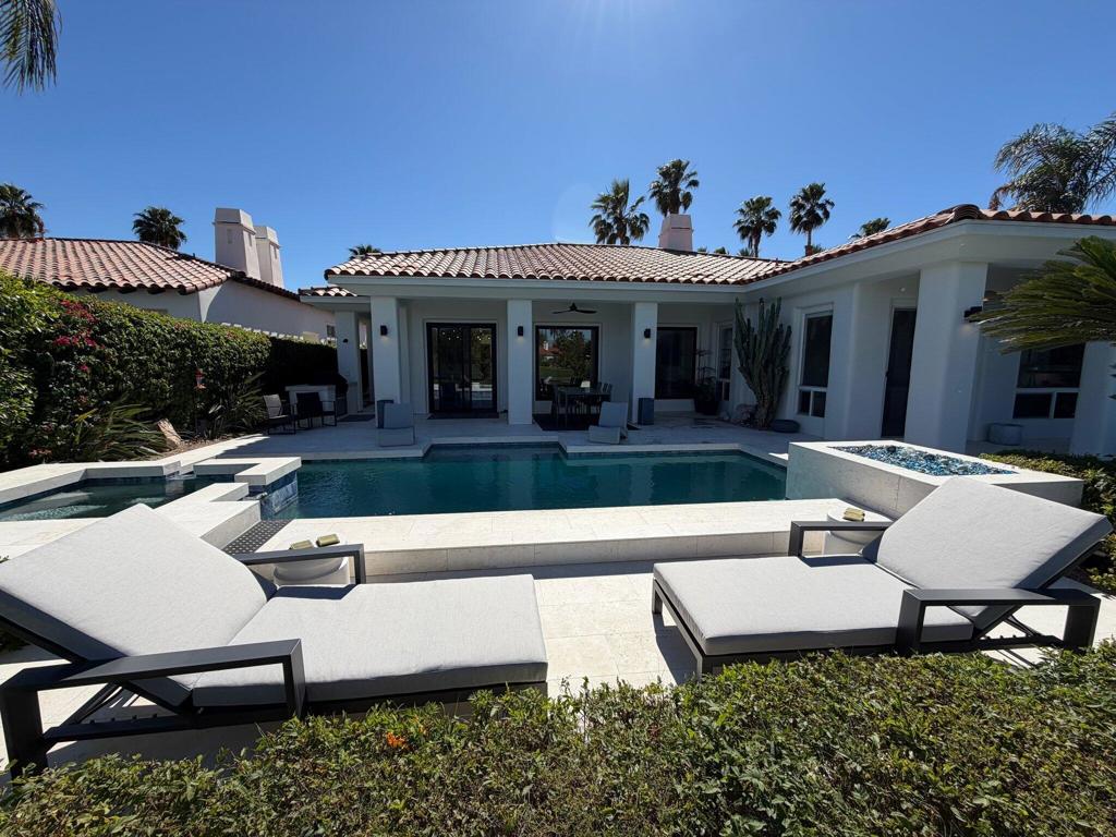 Single-story home with a turquoise pool, white patio loungers, and palm trees.