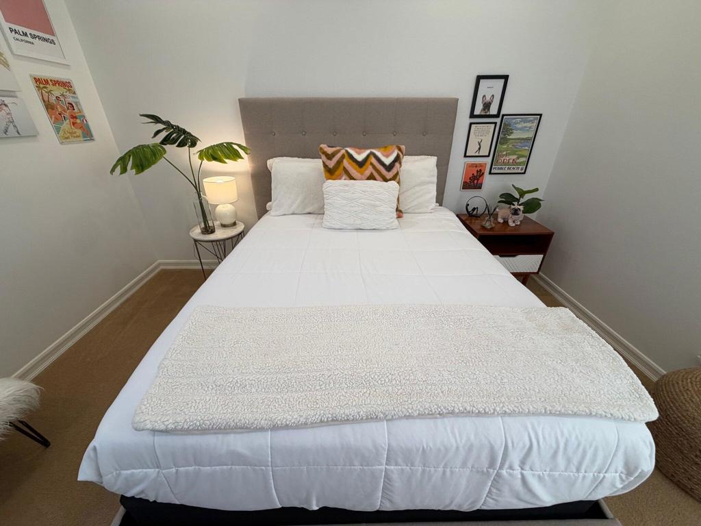 Staged bedroom with a white bed, tufted gray headboard, colorful accent pillows, and framed wall art.