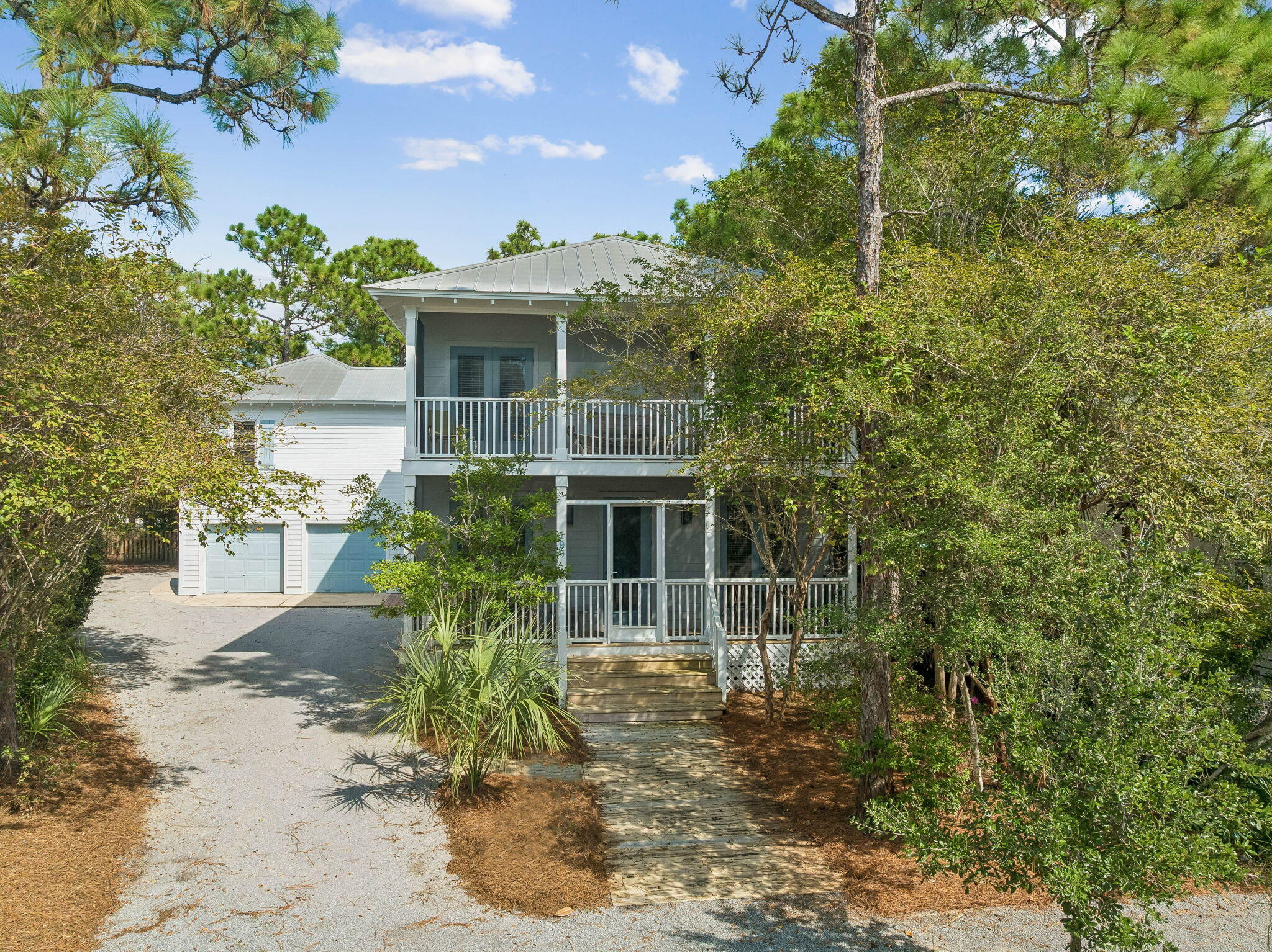Two-story gray coastal home with white porches and garage, framed by trees beside a gravel driveway.