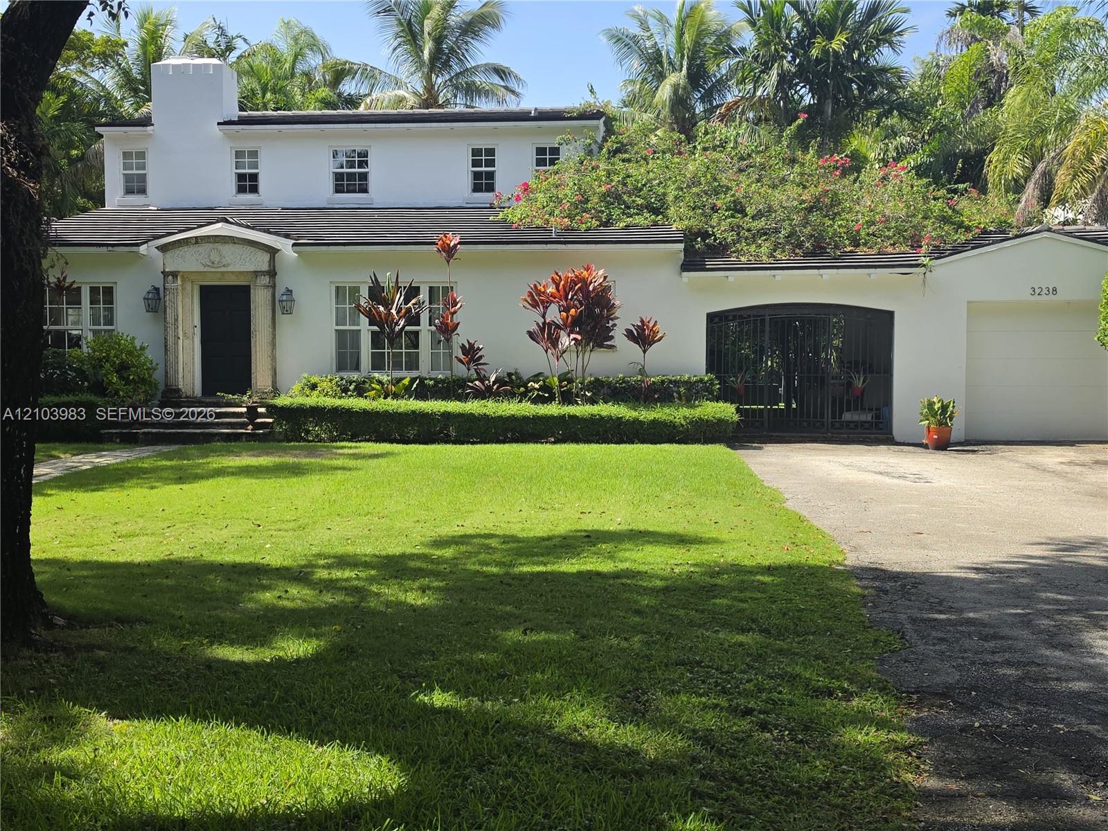 White two-story home with a columned entrance, gated driveway, and tropical landscaping.