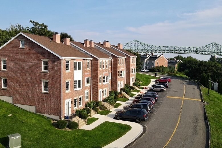 Red-brick townhouses with landscaped lawns and parked cars, with a steel bridge in the background.