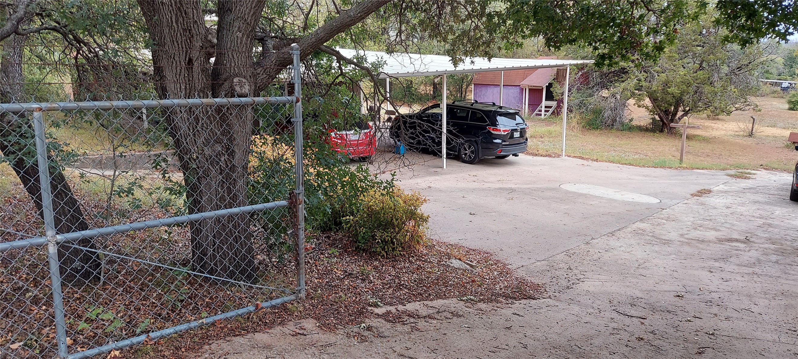 Concrete driveway and carport behind a chain-link fence, shaded by mature trees.