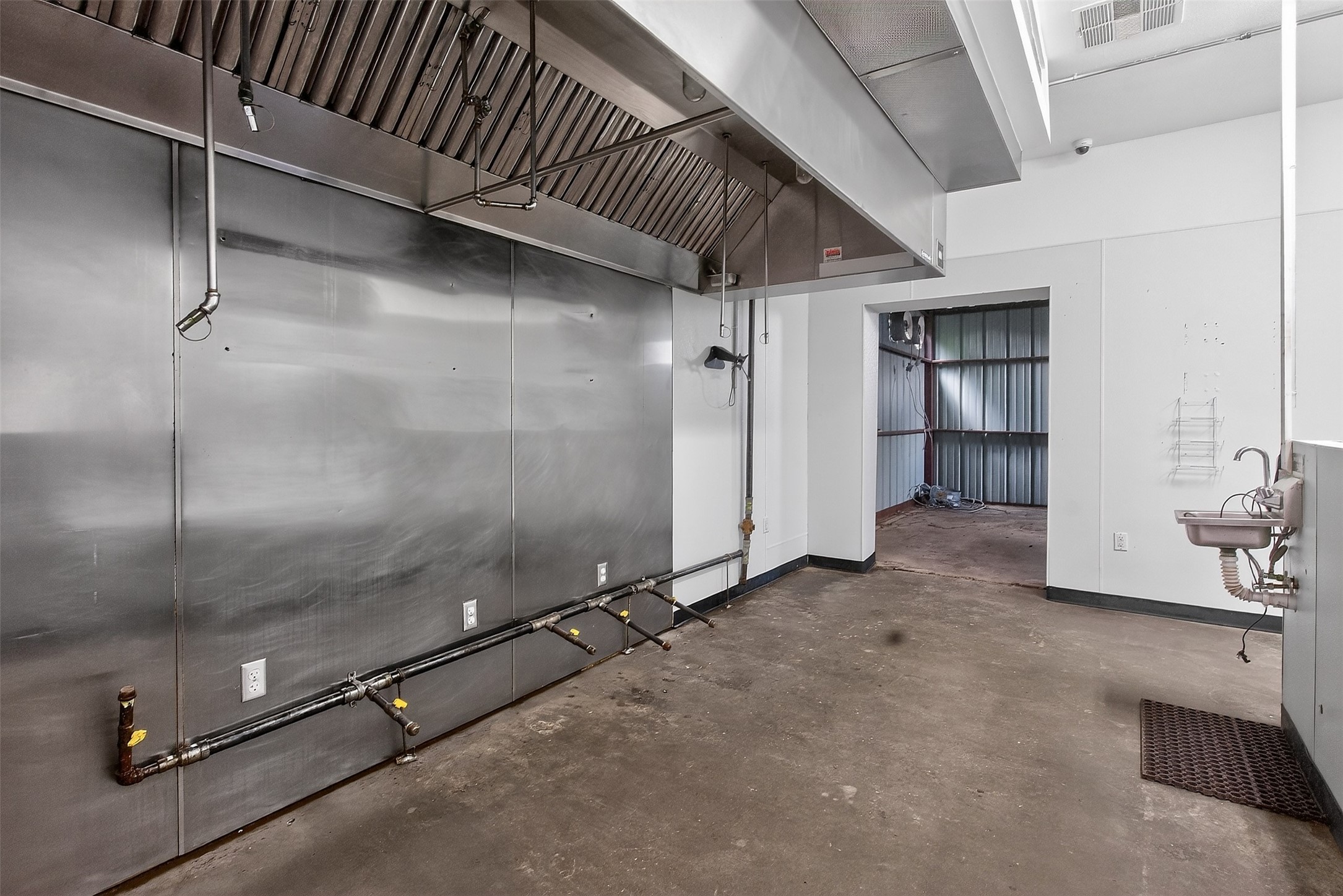 Commercial kitchen with a large stainless exhaust hood, exposed utility pipes, and a wall-mounted sink.