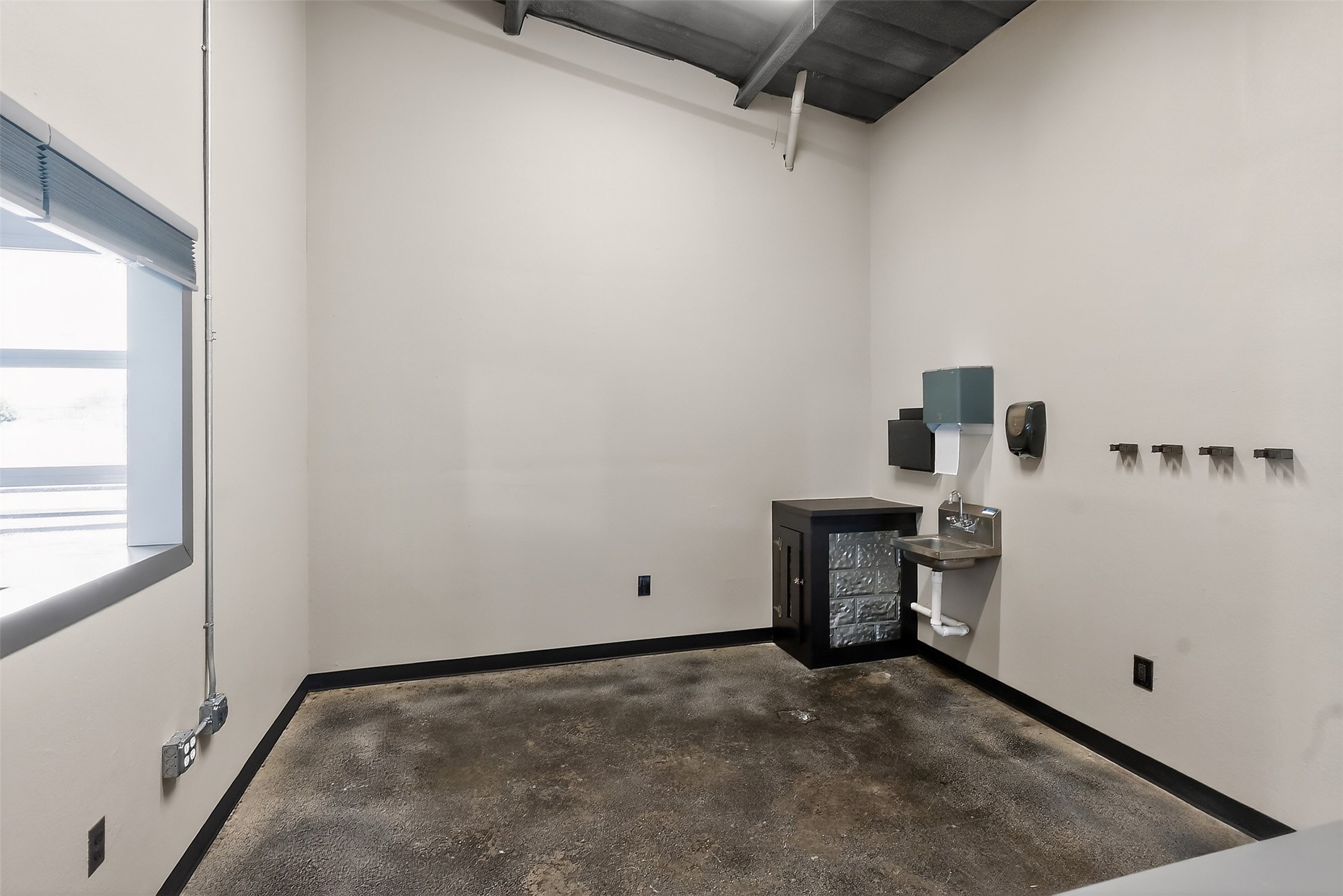 Utility room with a concrete floor, exposed ceiling, large window, and wall-mounted sink and dispensers.