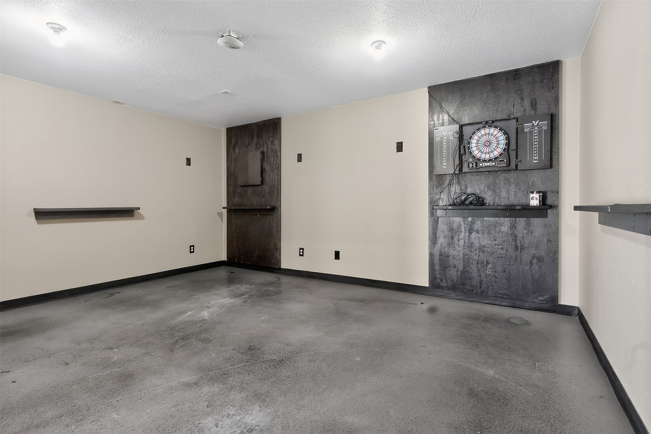 Empty room with a concrete floor and an electronic dartboard on a dark accent wall.