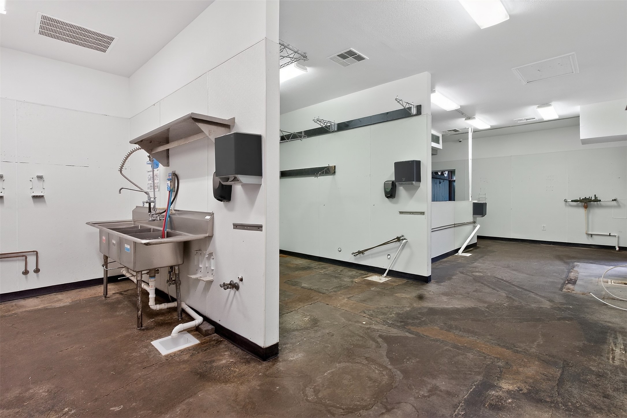 Vacant commercial room with a stainless-steel utility sink, exposed plumbing, and a worn concrete floor.