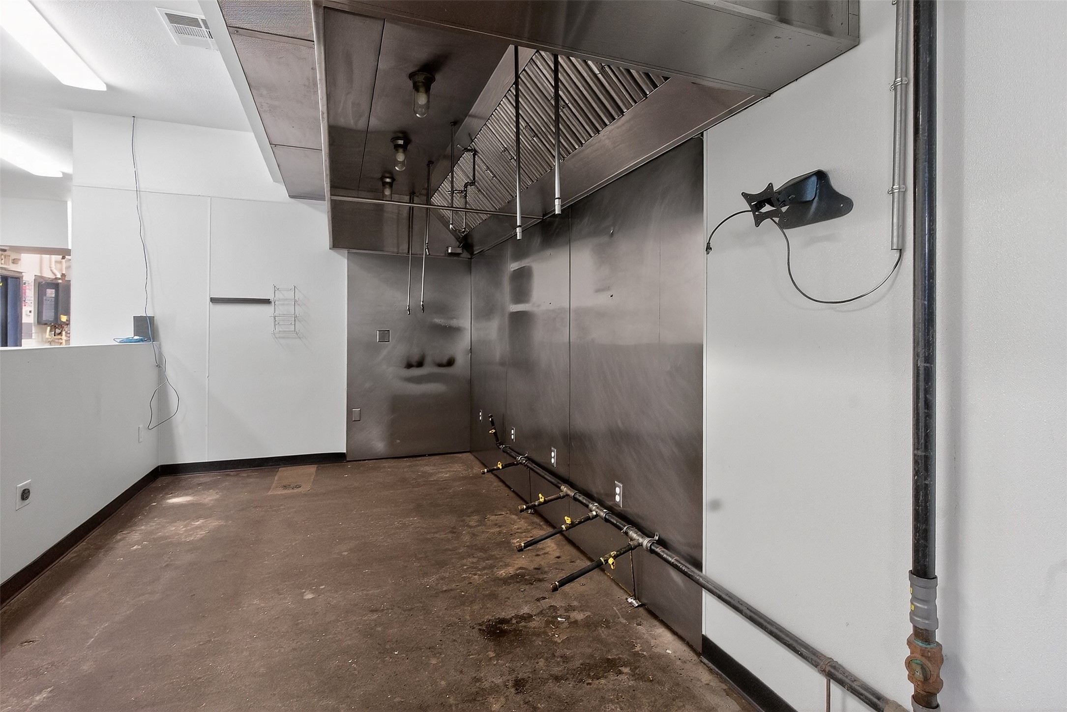 Empty commercial kitchen with a large stainless-steel exhaust hood and exposed piping along the wall.