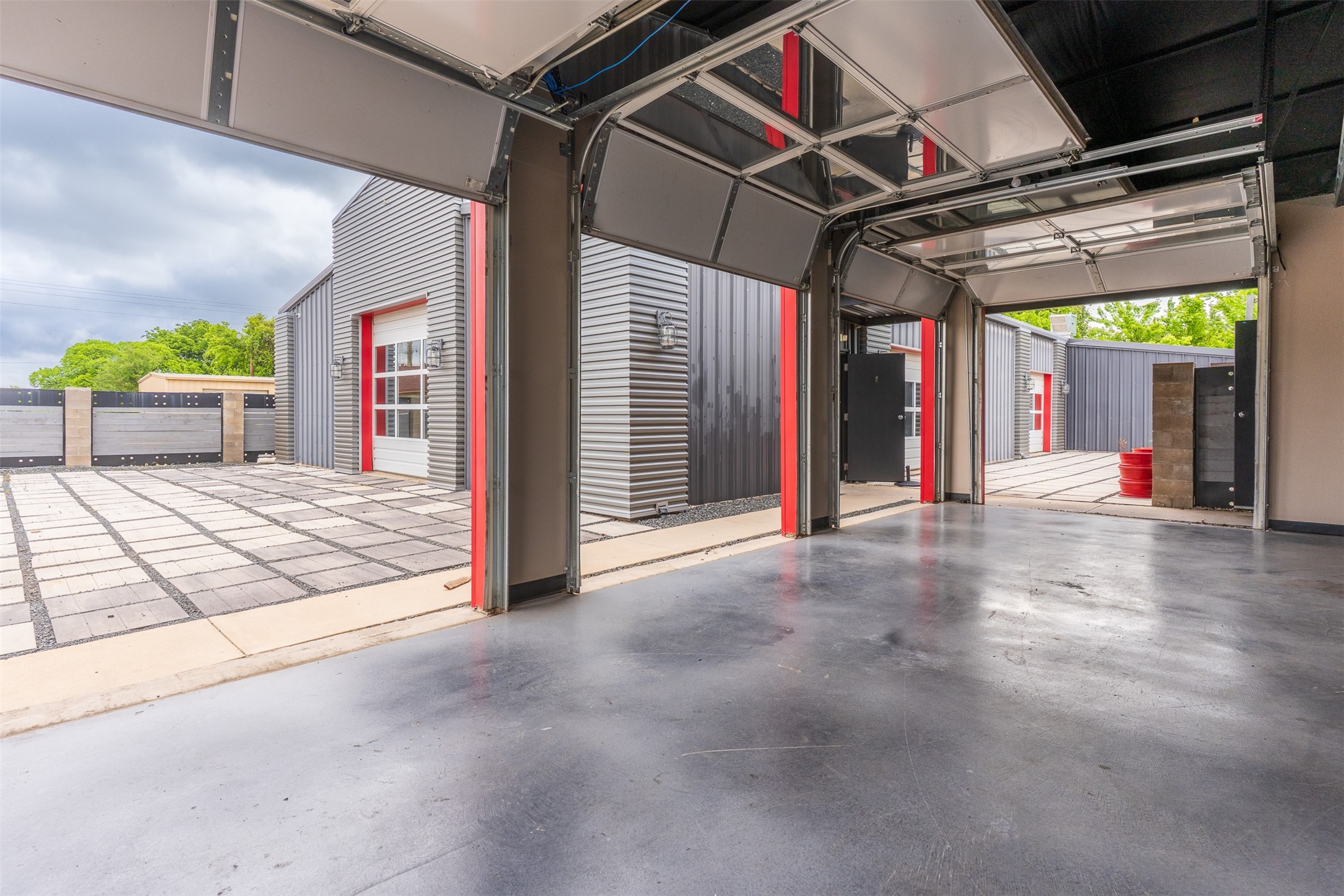 Open garage bay overlooking a paved courtyard and gray metal buildings with red trim.