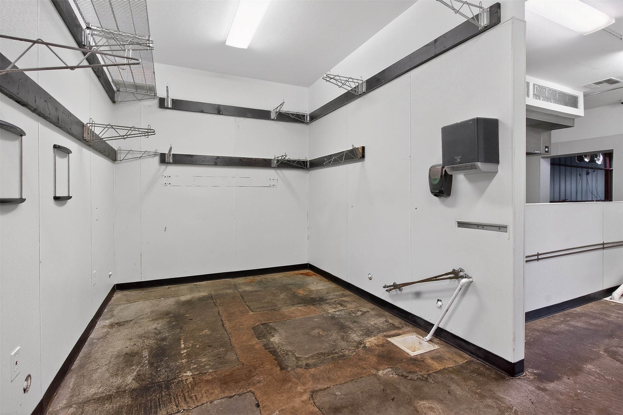 Empty commercial utility room with overhead racks, wall-mounted fixtures, and a worn concrete floor.