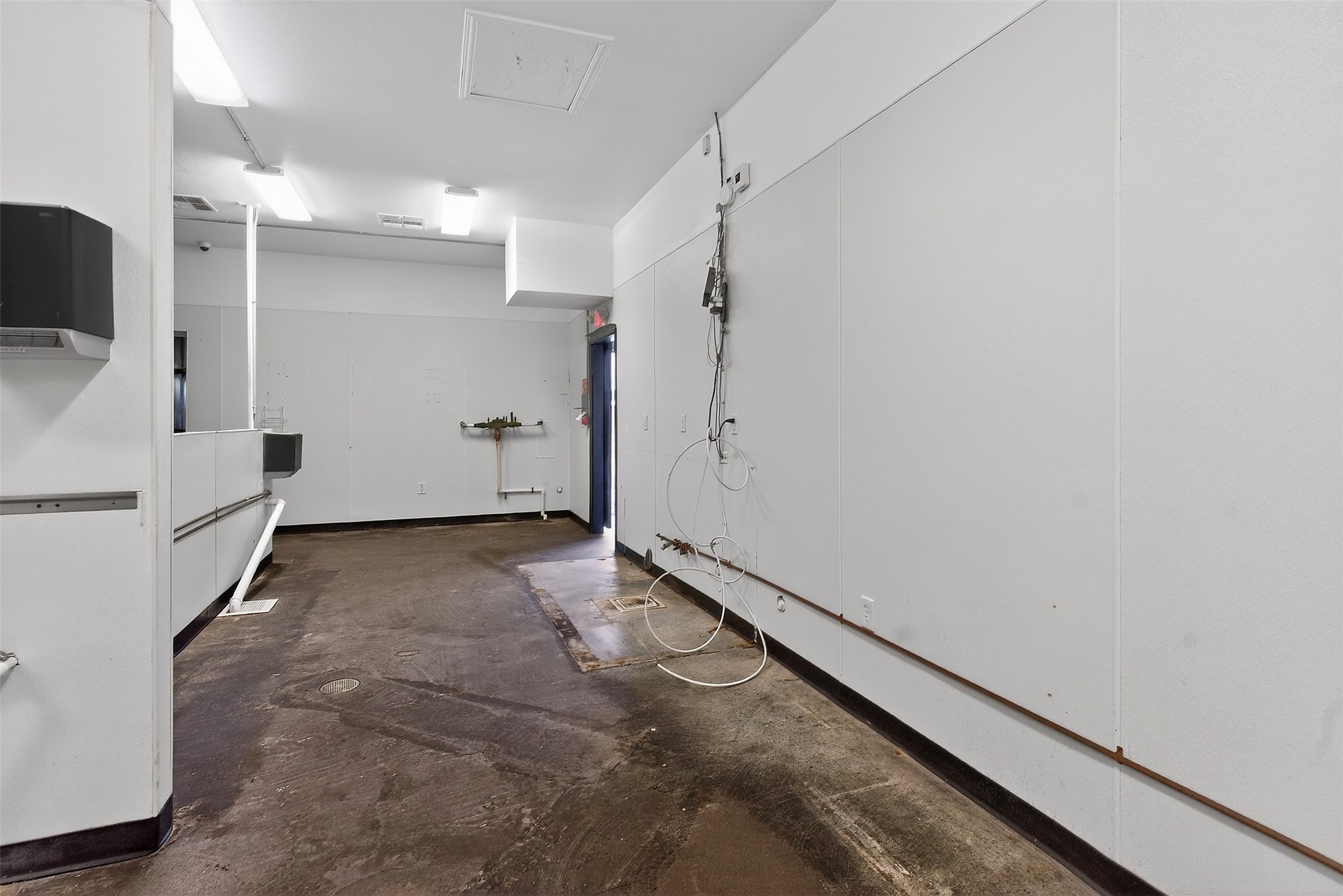 Empty commercial workroom with a worn concrete floor, floor drains, and exposed plumbing.