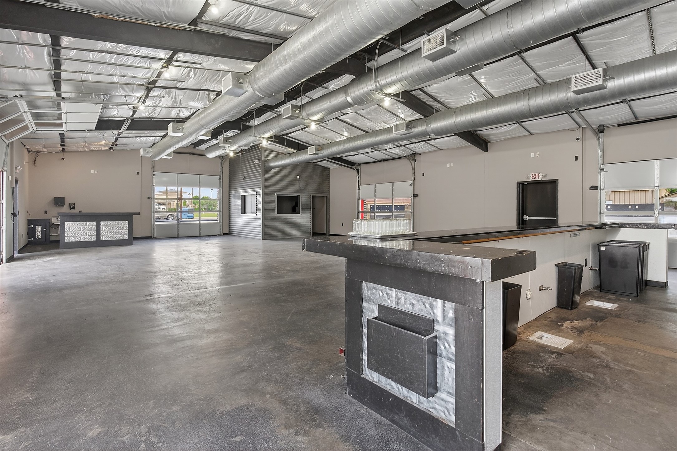 Open commercial interior with overhead garage doors, exposed ductwork, and a long black counter.