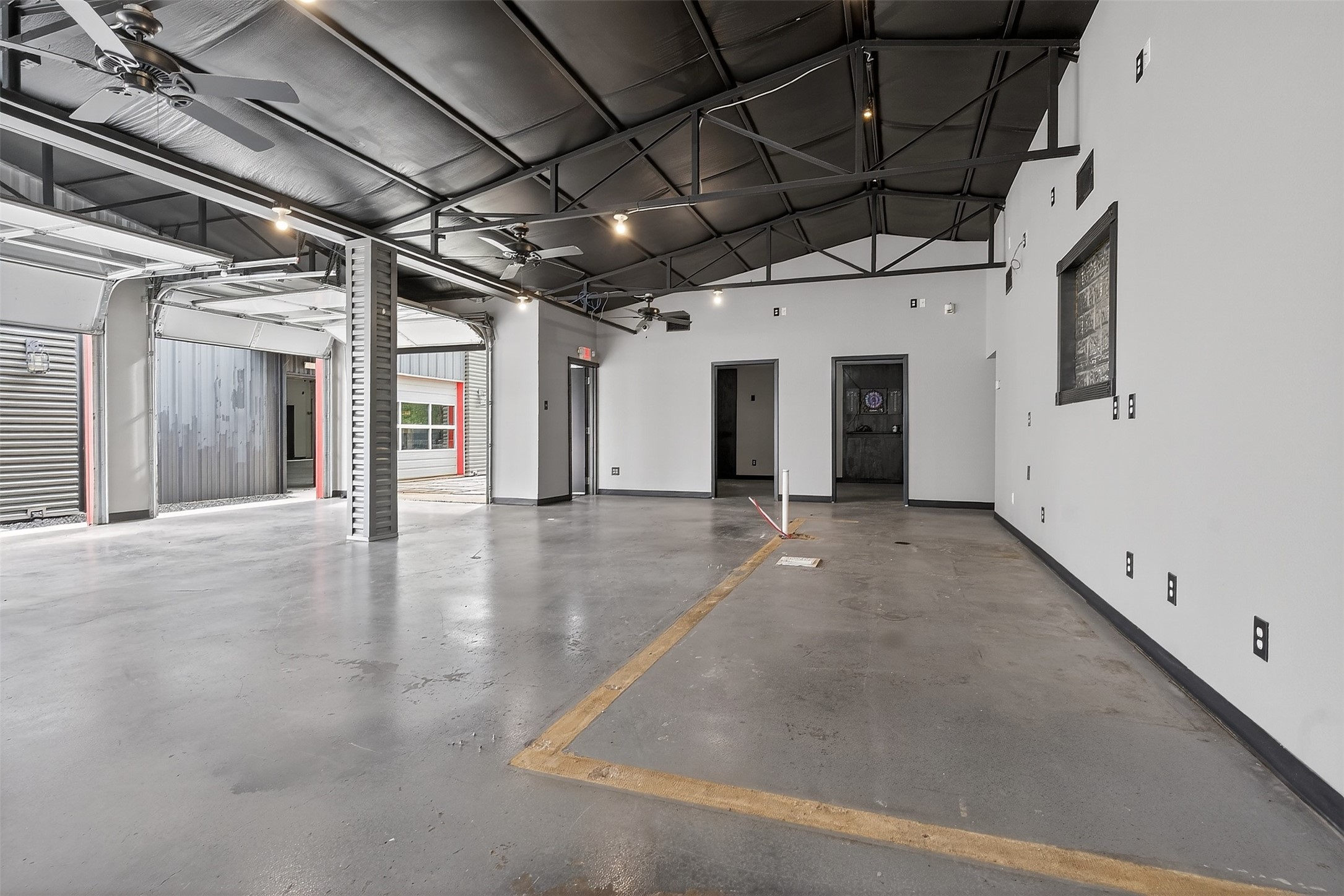 Vacant industrial garage with open vehicle bays, exposed roof trusses, ceiling fans, and a marked concrete floor.