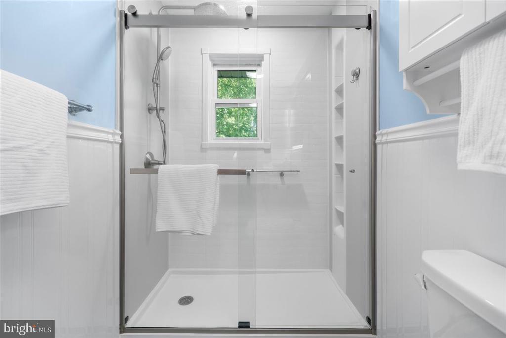 Bathroom with a white tiled walk-in shower, clear sliding door, built-in shelves, and a window overlooking greenery.