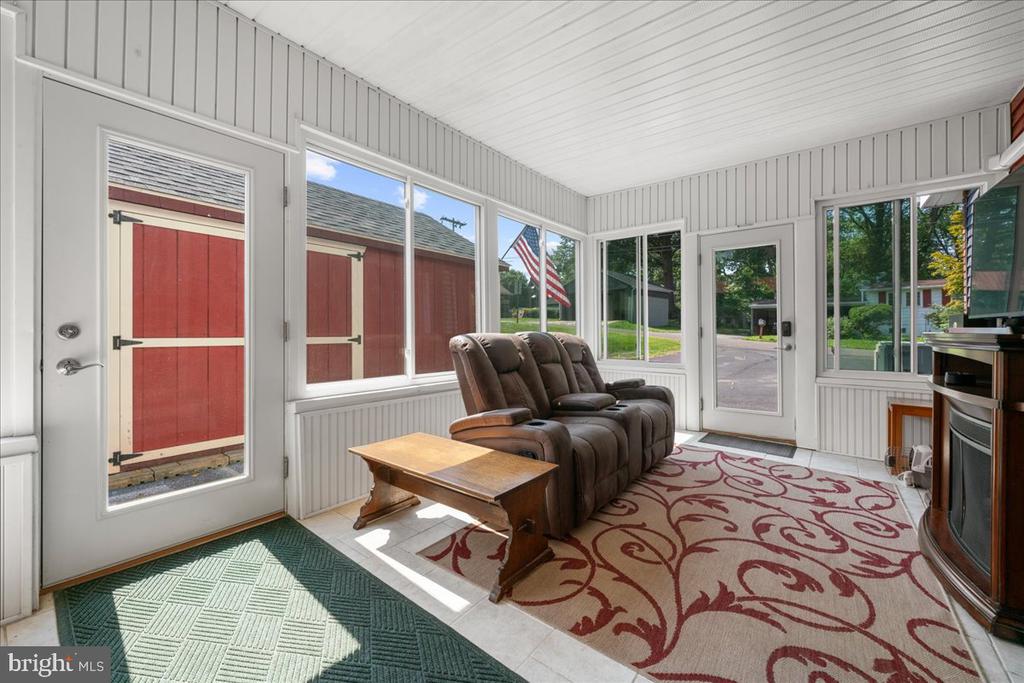 Enclosed sunroom with reclining chairs, patterned rugs, and large windows overlooking a red outbuilding.