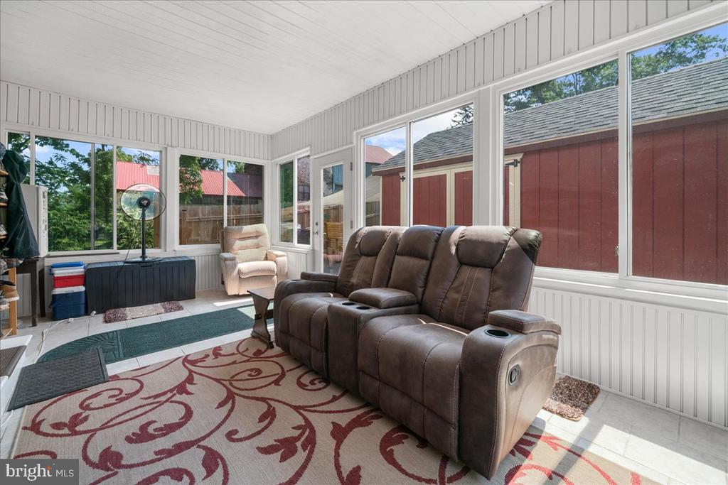 Enclosed sunroom with a brown reclining sofa, an armchair, and large windows overlooking a red outbuilding.