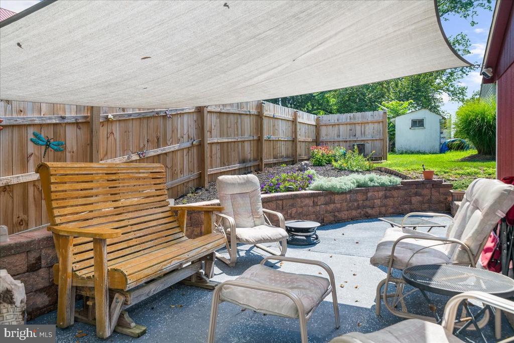 Backyard patio with a shade sail, wooden bench, cushioned chairs, raised flower beds, and fenced yard.