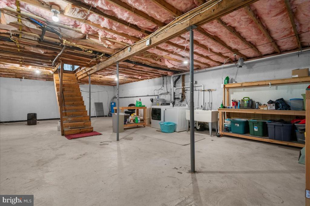 Unfinished basement with exposed ceiling insulation, wooden stairs, laundry appliances, and storage shelves.