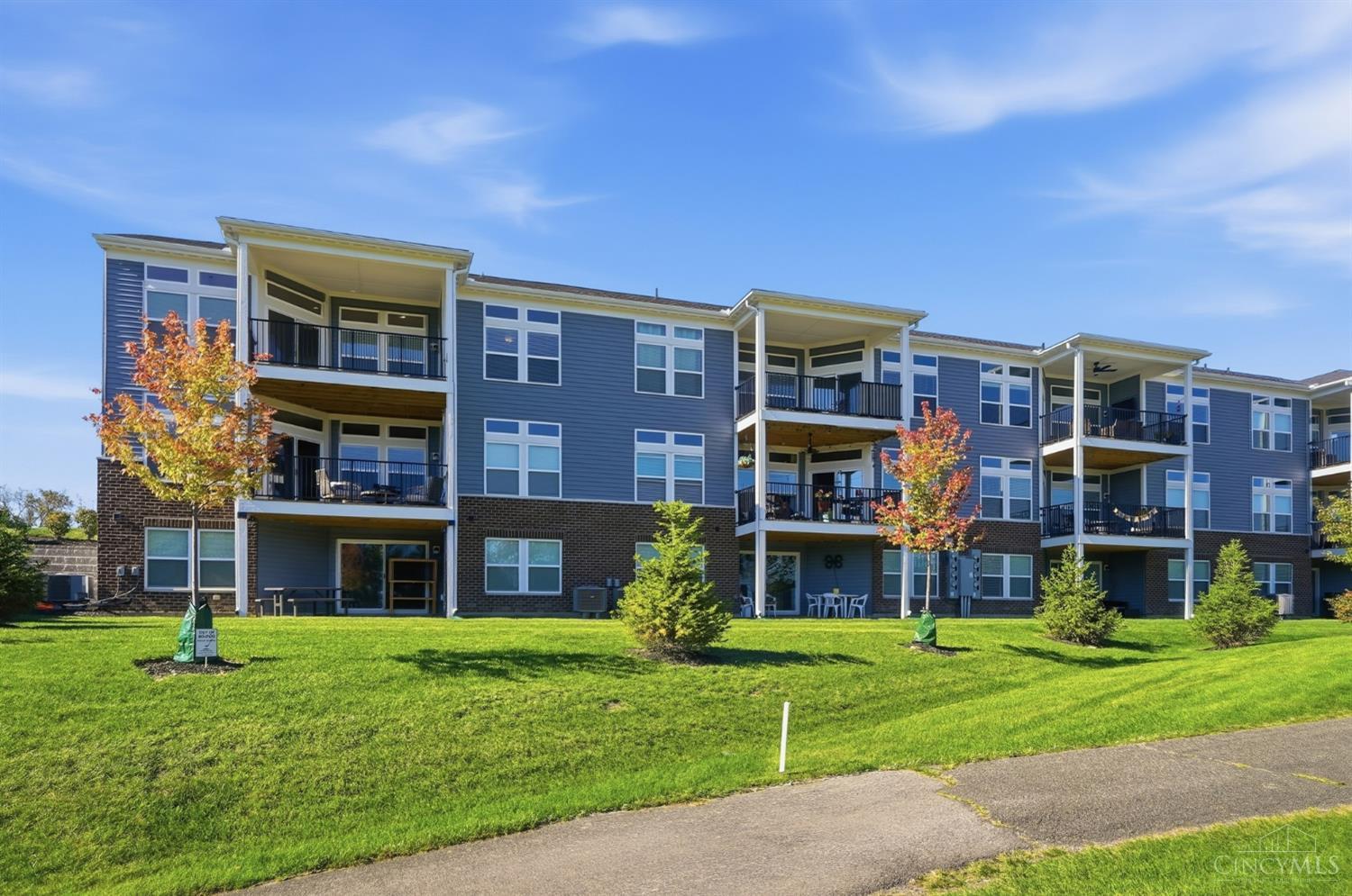Three-story blue-gray apartment building with balconies, brick ground level, and autumn trees along a green lawn.