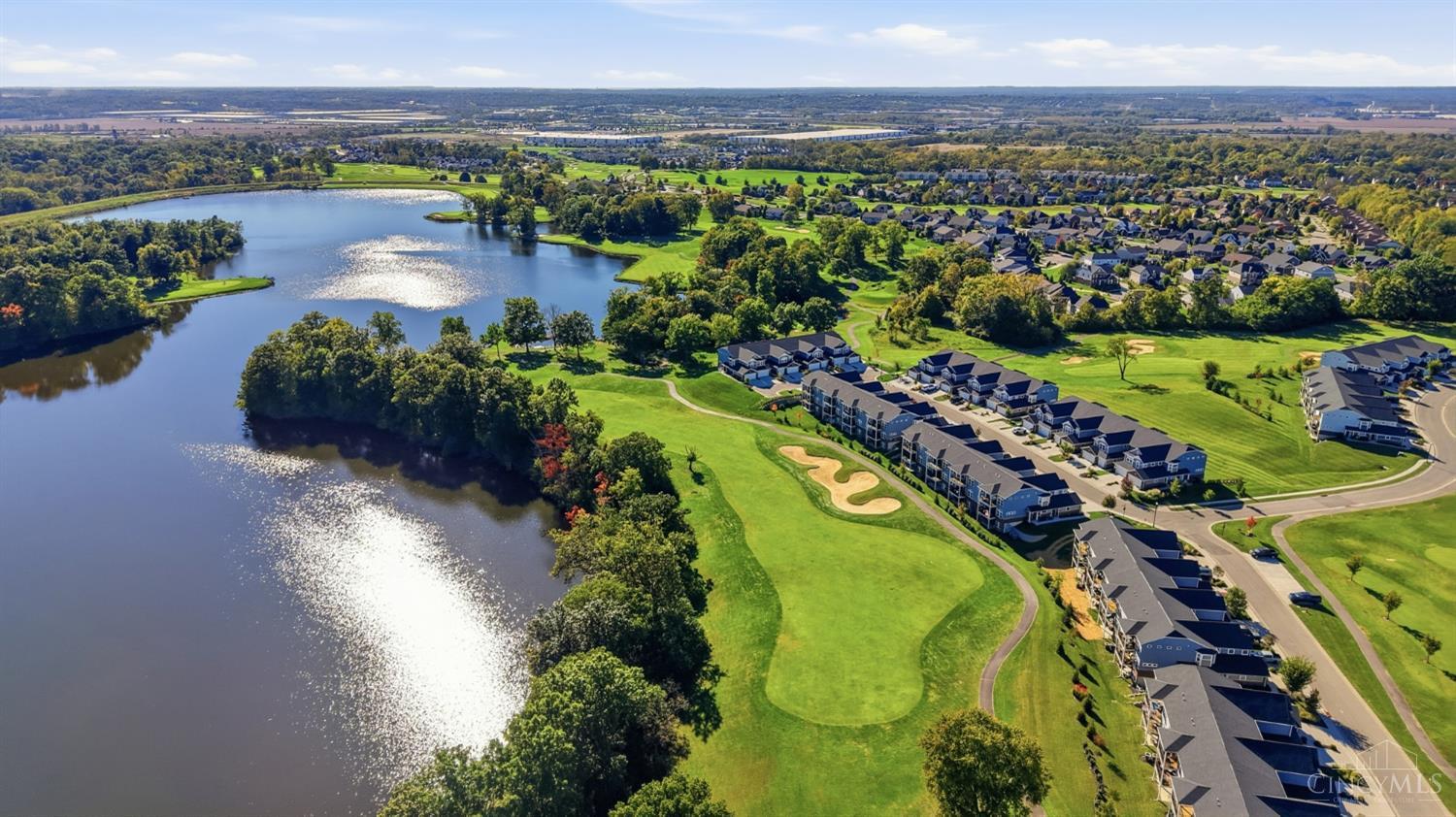 Aerial view of homes bordering a lake and golf course with a winding fairway and sand bunker.