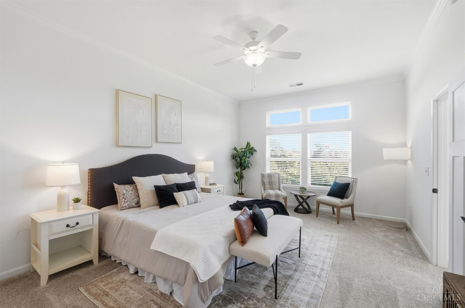 Staged bedroom with a large bed, dark upholstered headboard, and a sitting area beside wide windows.