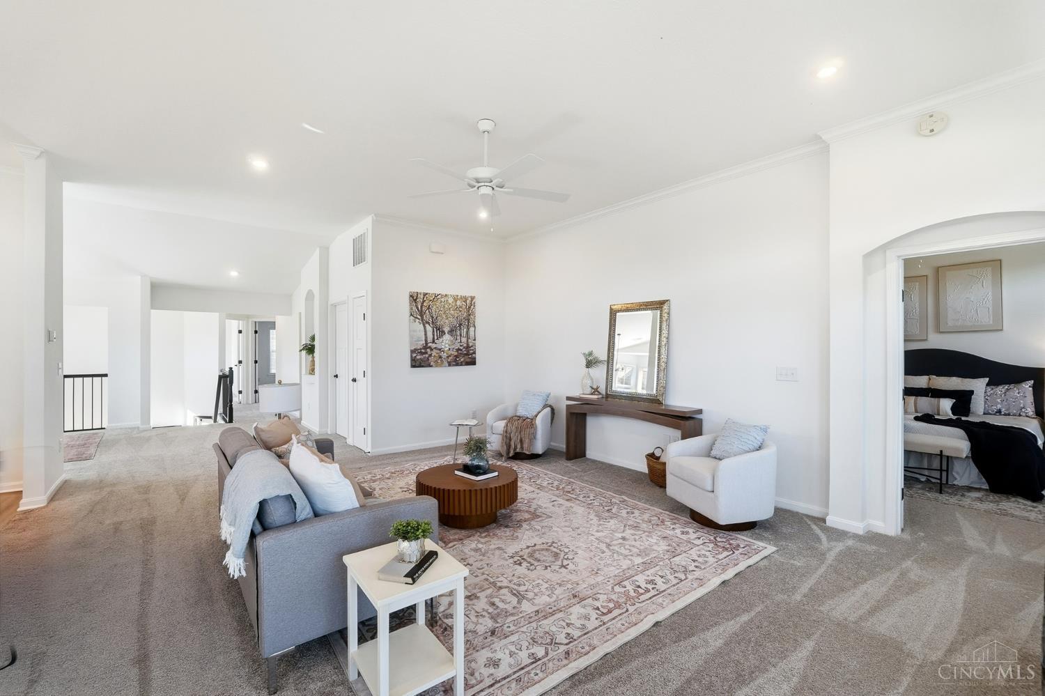 Staged living room with a patterned rug, gray sofa, white armchairs, and a bedroom visible through the doorway.