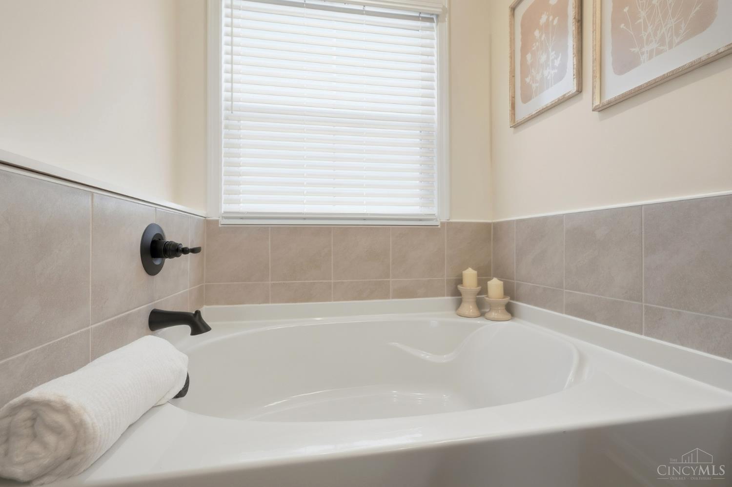 White soaking tub with beige tile, black fixtures, and a window with white blinds.