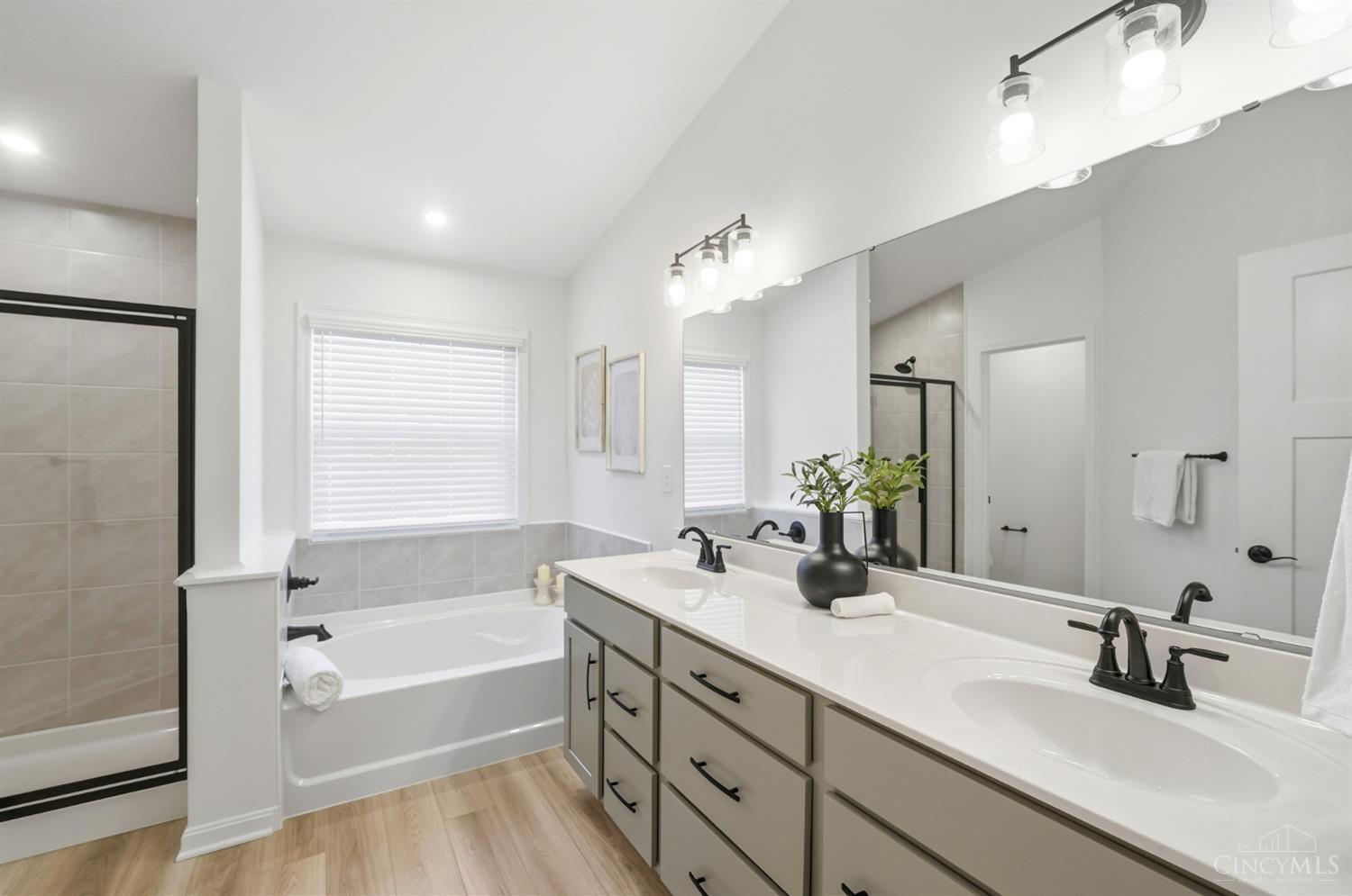 Bright bathroom with a long double vanity, bathtub beneath a window, and glass-enclosed shower.