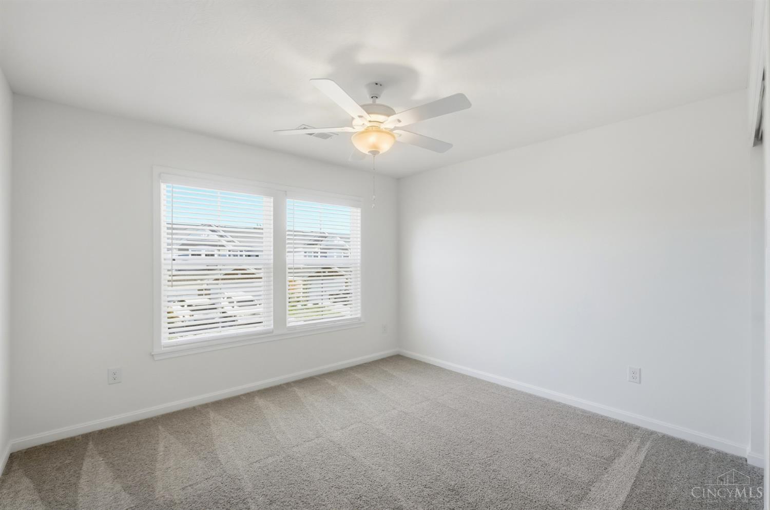 Empty bedroom with gray carpet, two blind-covered windows, and a ceiling fan.