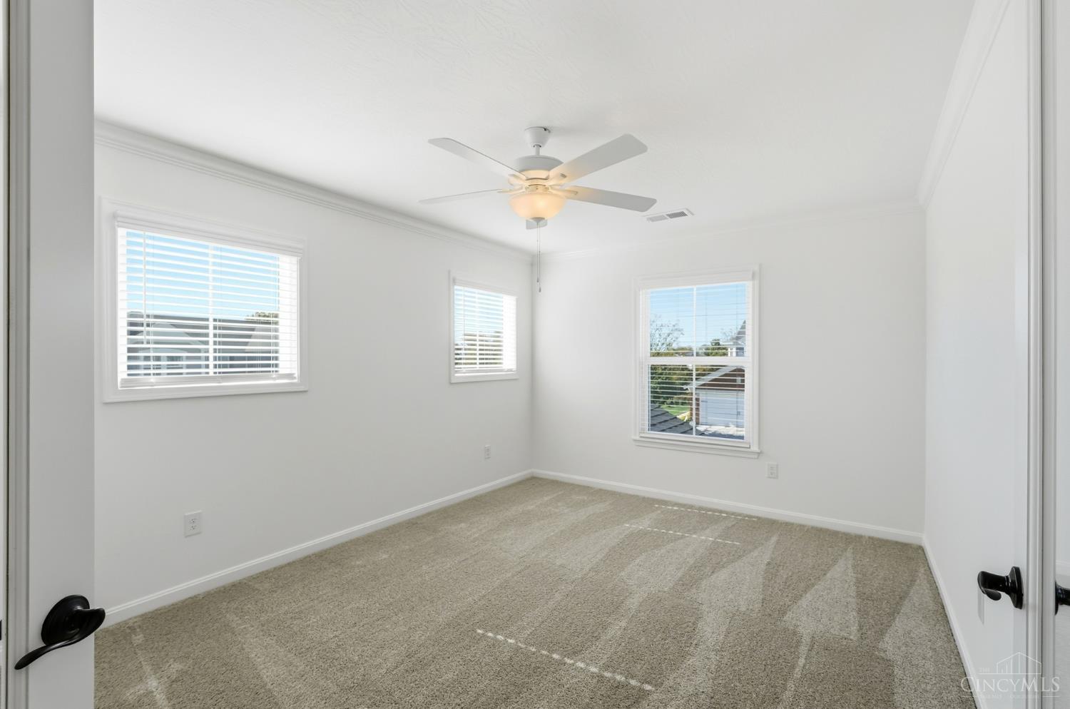 Empty bedroom with beige carpet, three windows, and a ceiling fan with light.