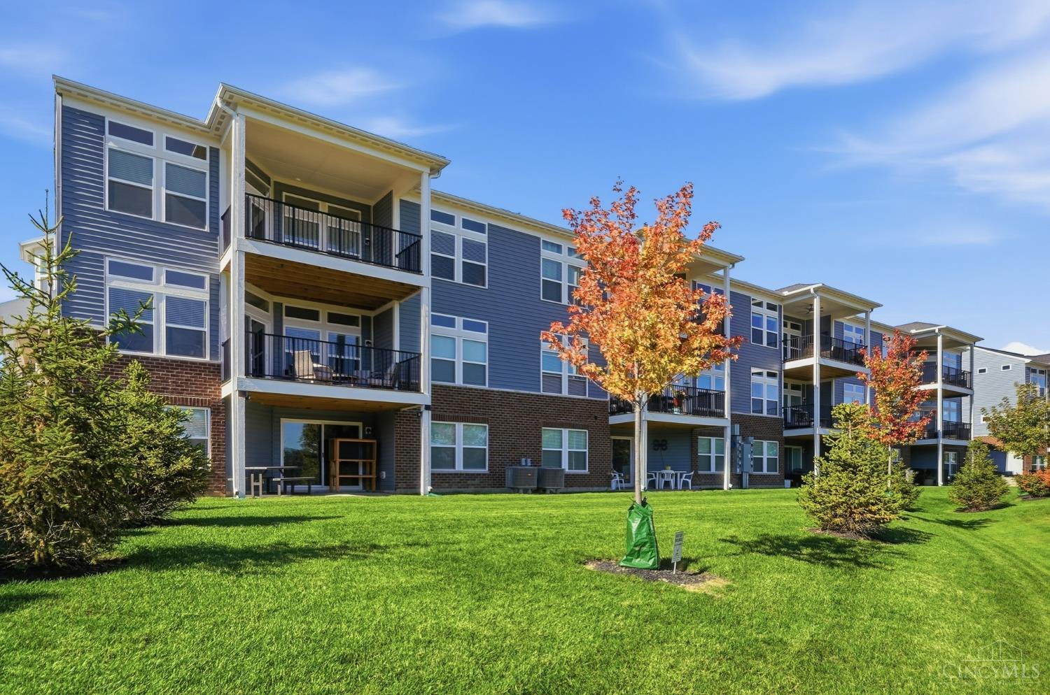 Blue-gray residential building with stacked balconies, brick lower walls, and autumn trees along a green lawn.