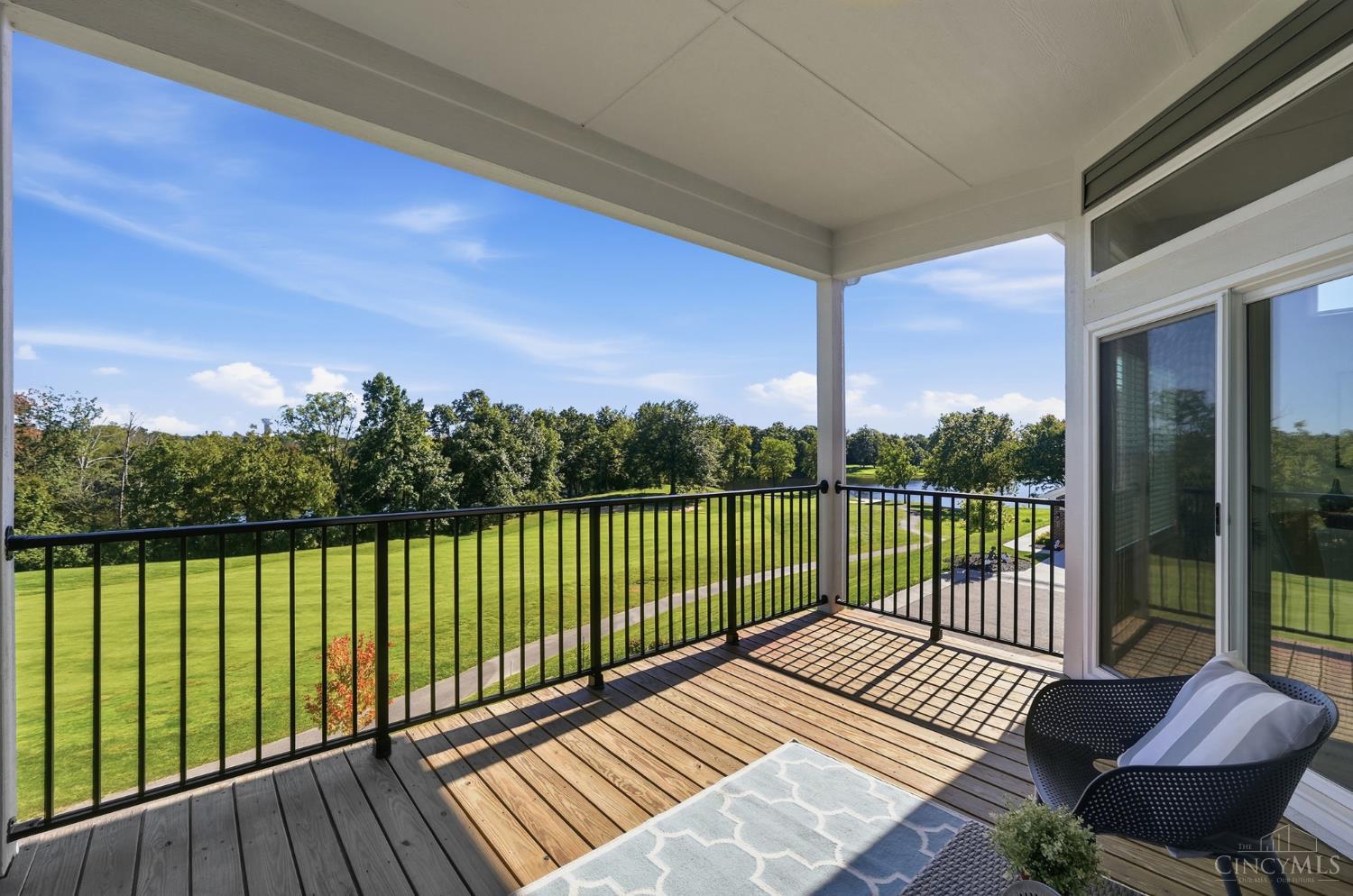 Covered balcony with wood decking and black railing overlooking a green lawn, trees, and water.